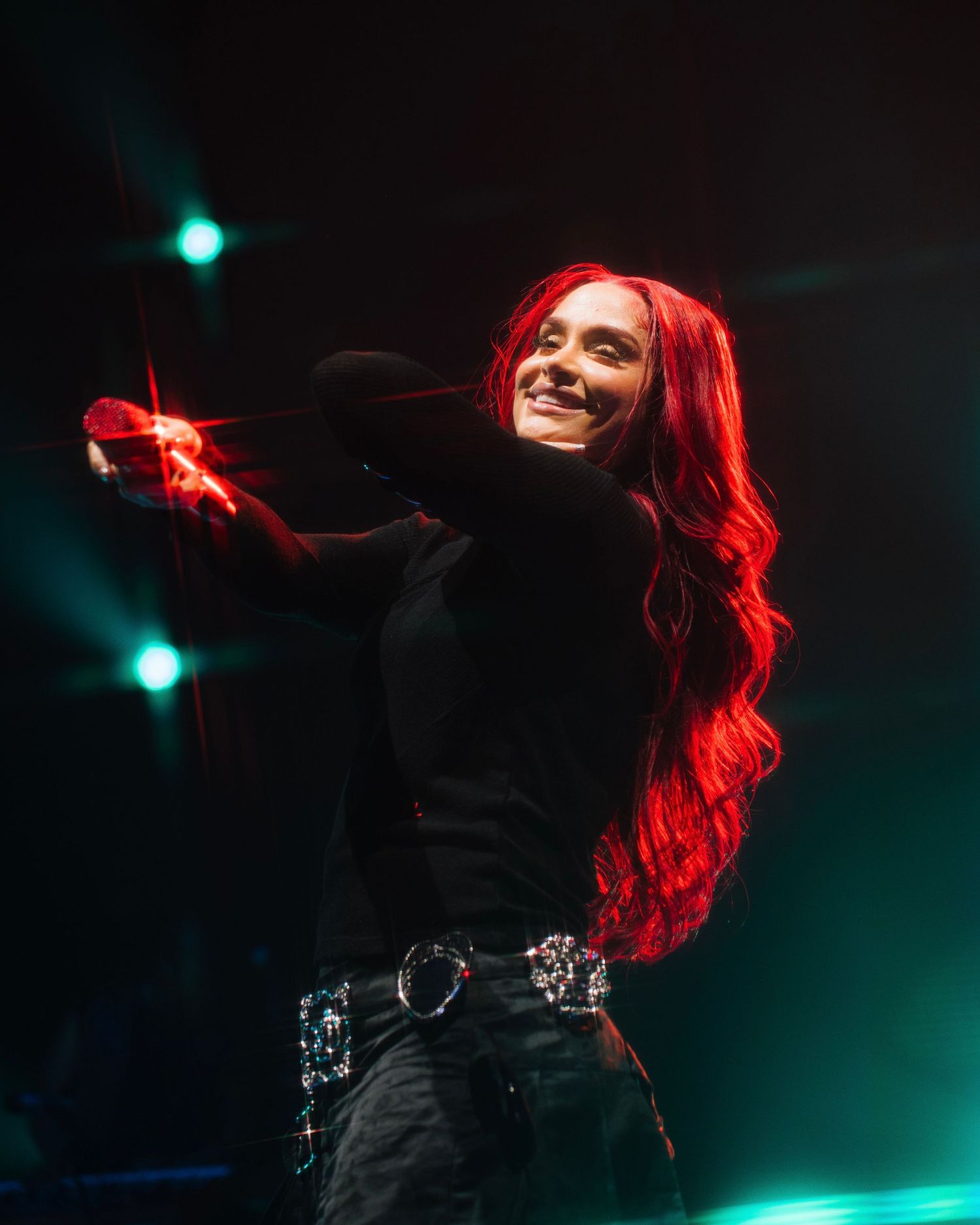 Performer with vibrant red hair in black outfit dances on stage with dramatic turquoise lighting effects.