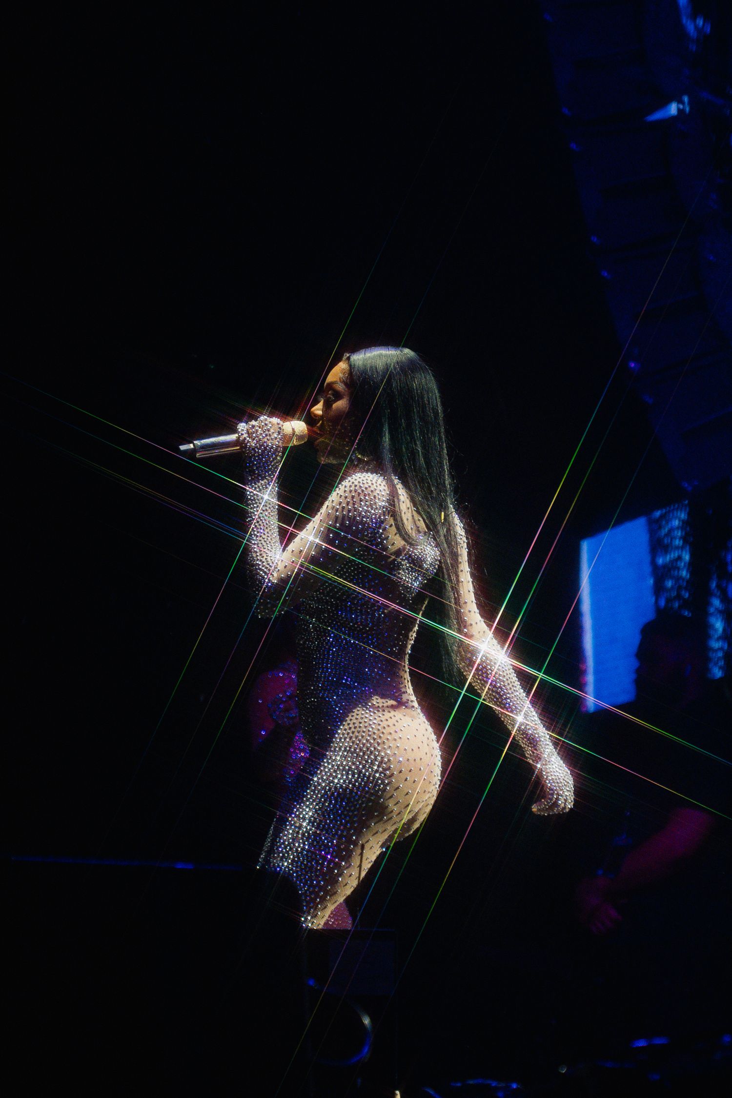 Singer performs on dark stage wearing a sparkling bodysuit with dramatic lighting effects creating starbursts around them.