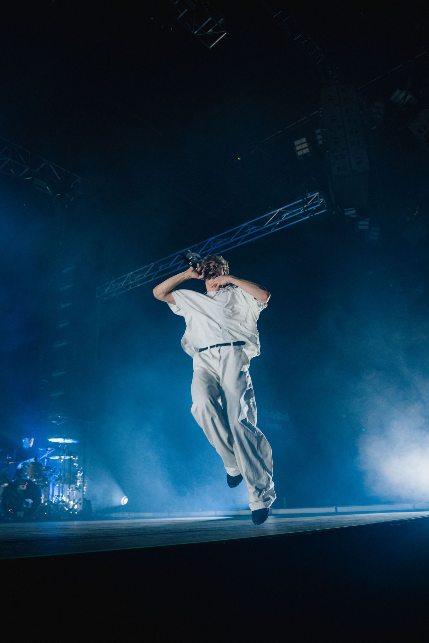 Performer in white outfit leaps high on a dark stage with dramatic blue lighting and fog effects during concert performance.