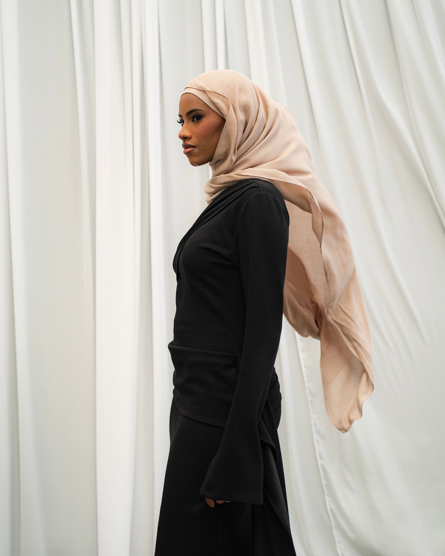 Elegant portrait of person in black outfit and beige hijab against white curtain backdrop.