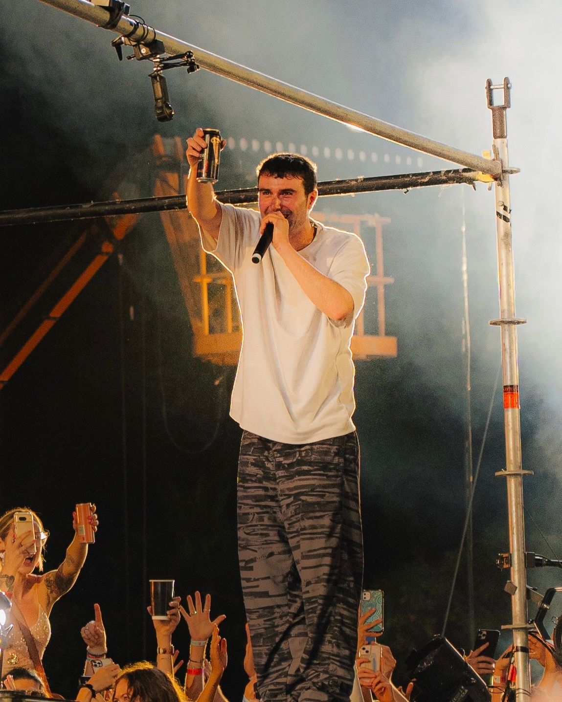Artist in white t-shirt performing on foggy stage with raised hands from crowd visible.