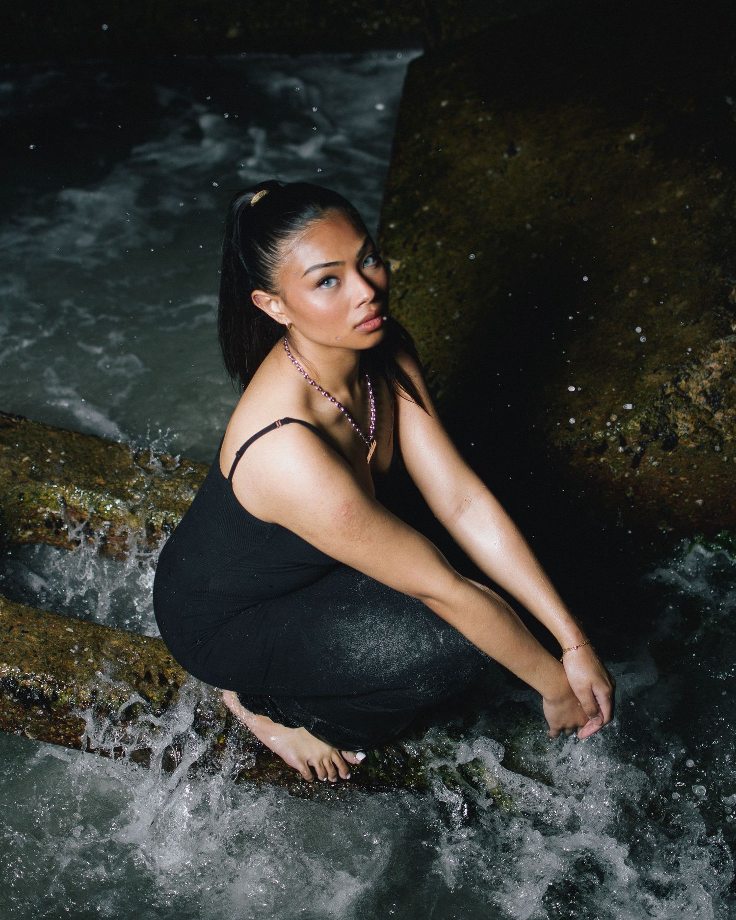 An artistic photo showing a figure in a black dress crouching near rushing water in a moody natural setting.