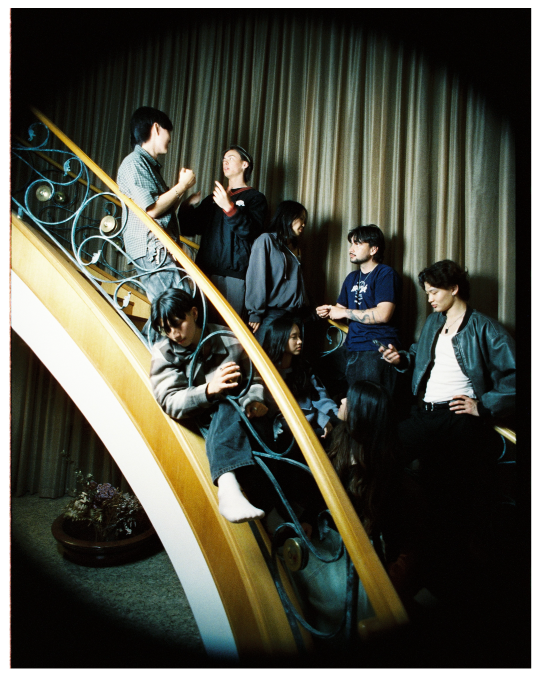 A group lounges on curved marble stairs with gold railings against a dark curtained backdrop in moody lighting.