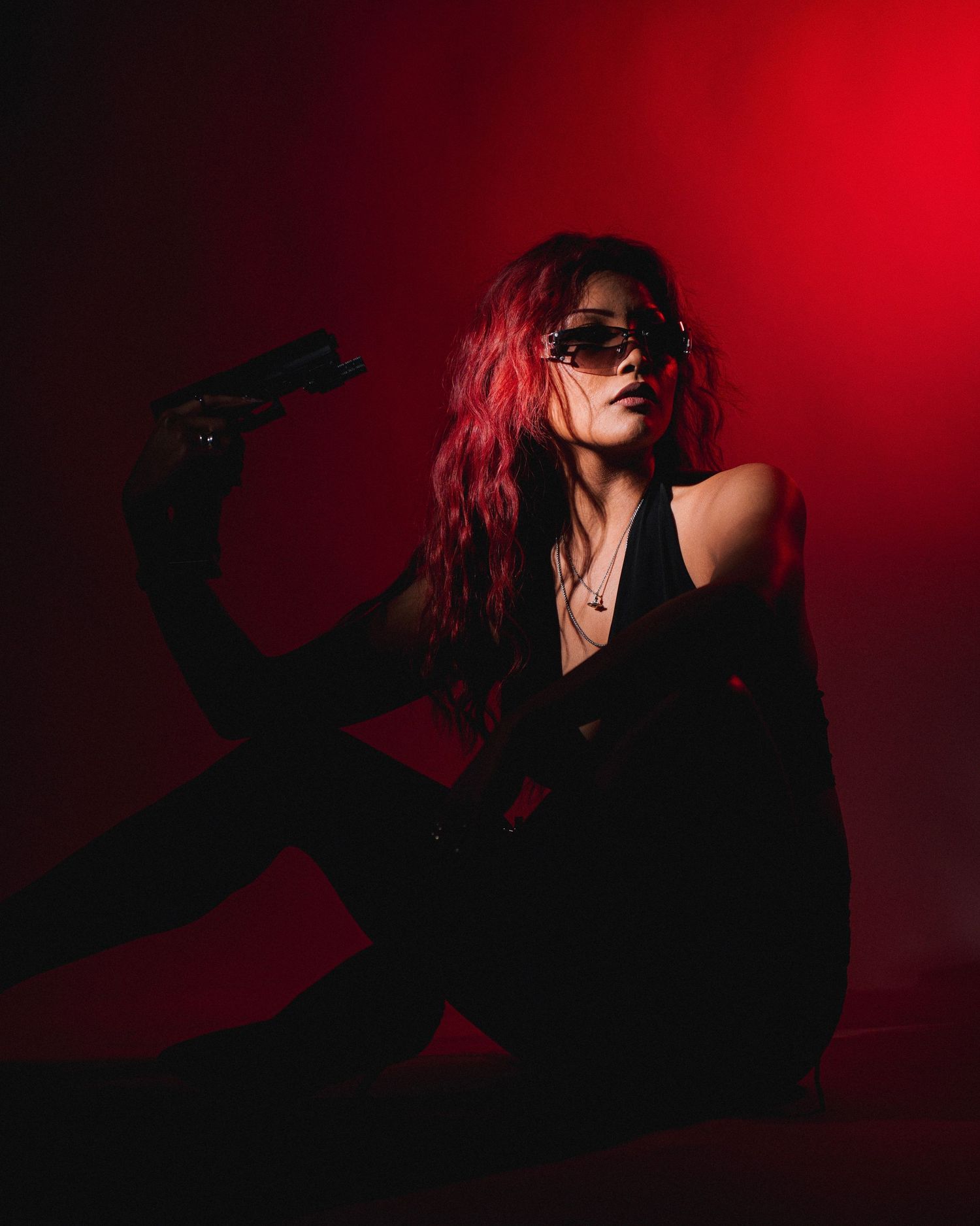 A figure in black clothing and sunglasses with pink hair poses dramatically against a dark red background in moody lighting.