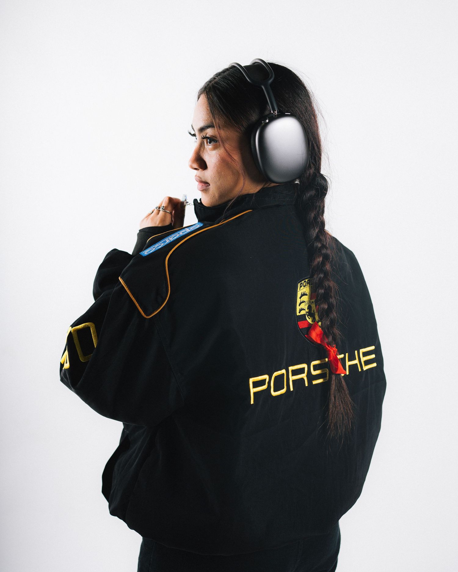 Person in black Porsche racing jacket with white headphones and braided hair against white background.
