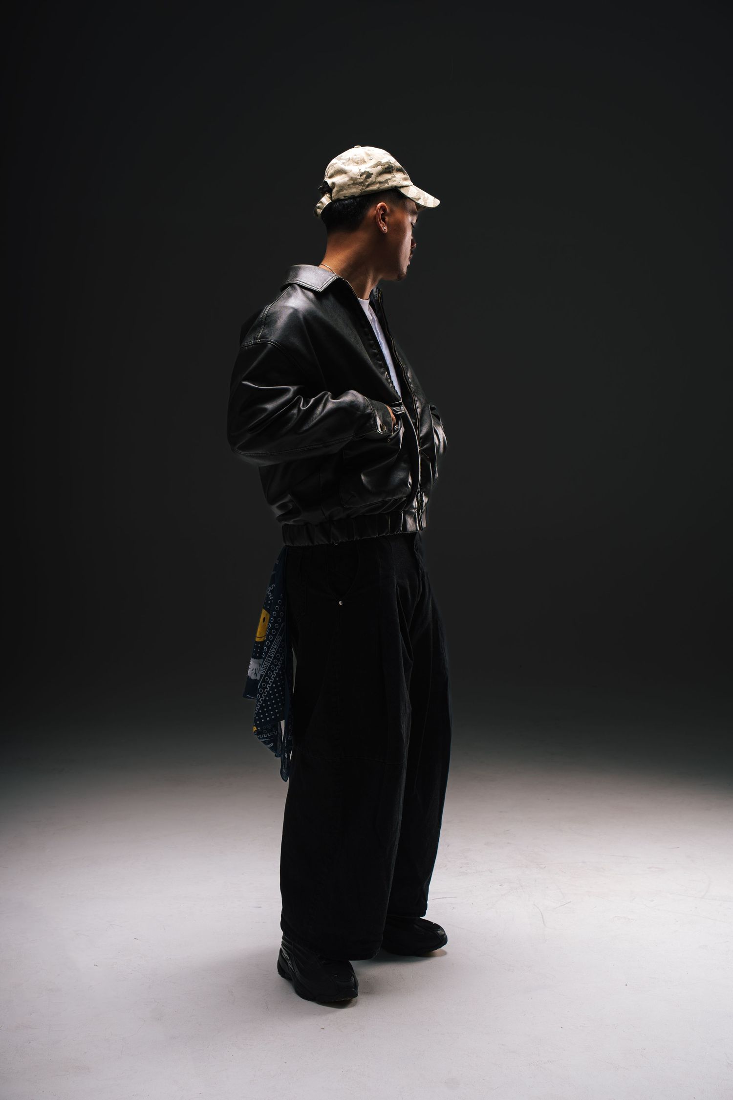Side view of person in black bomber jacket and loose pants with baseball cap against dark backdrop in studio lighting.