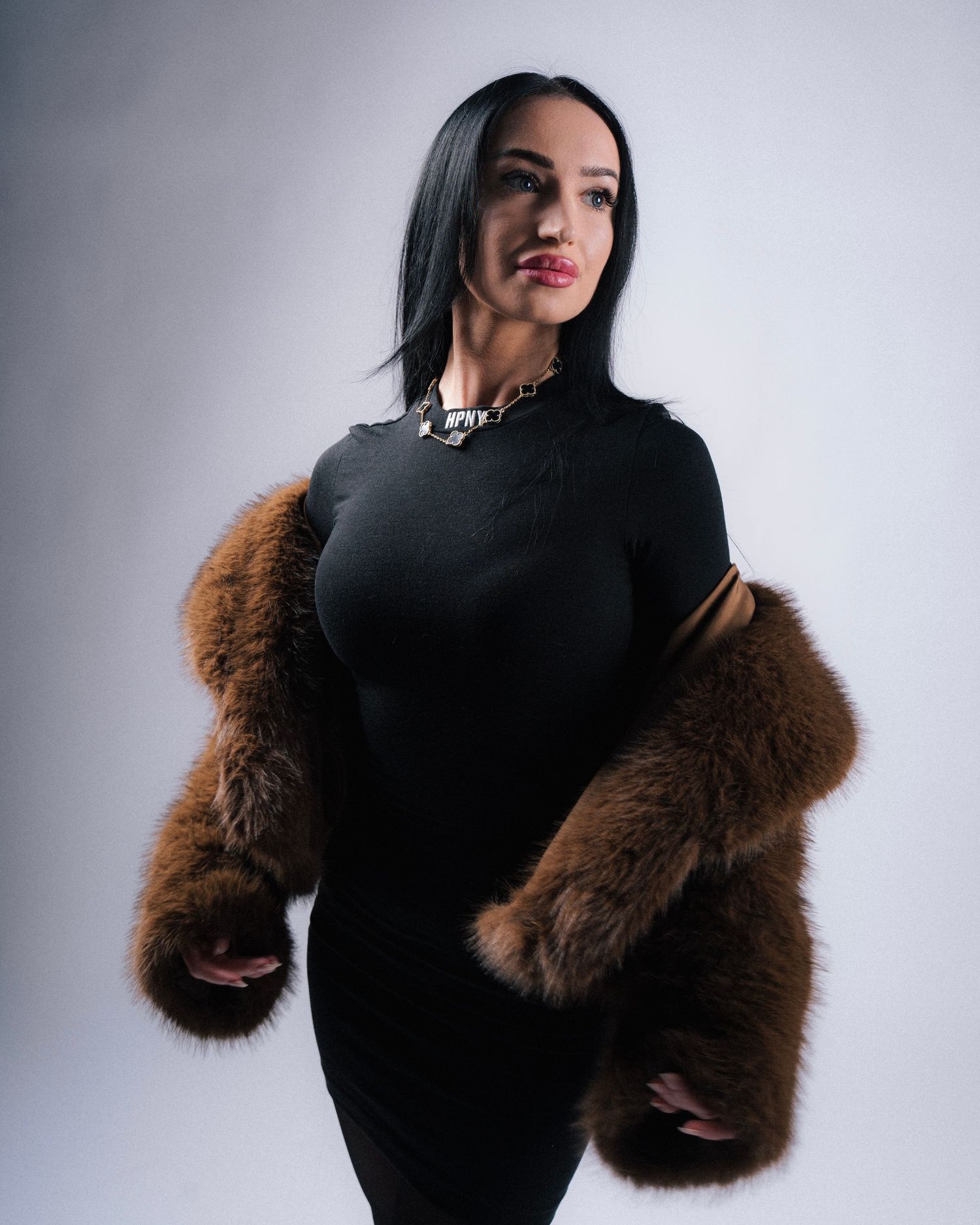 Fashion portrait in black bodycon dress with brown fur sleeve accents and silver chain necklace against light background.