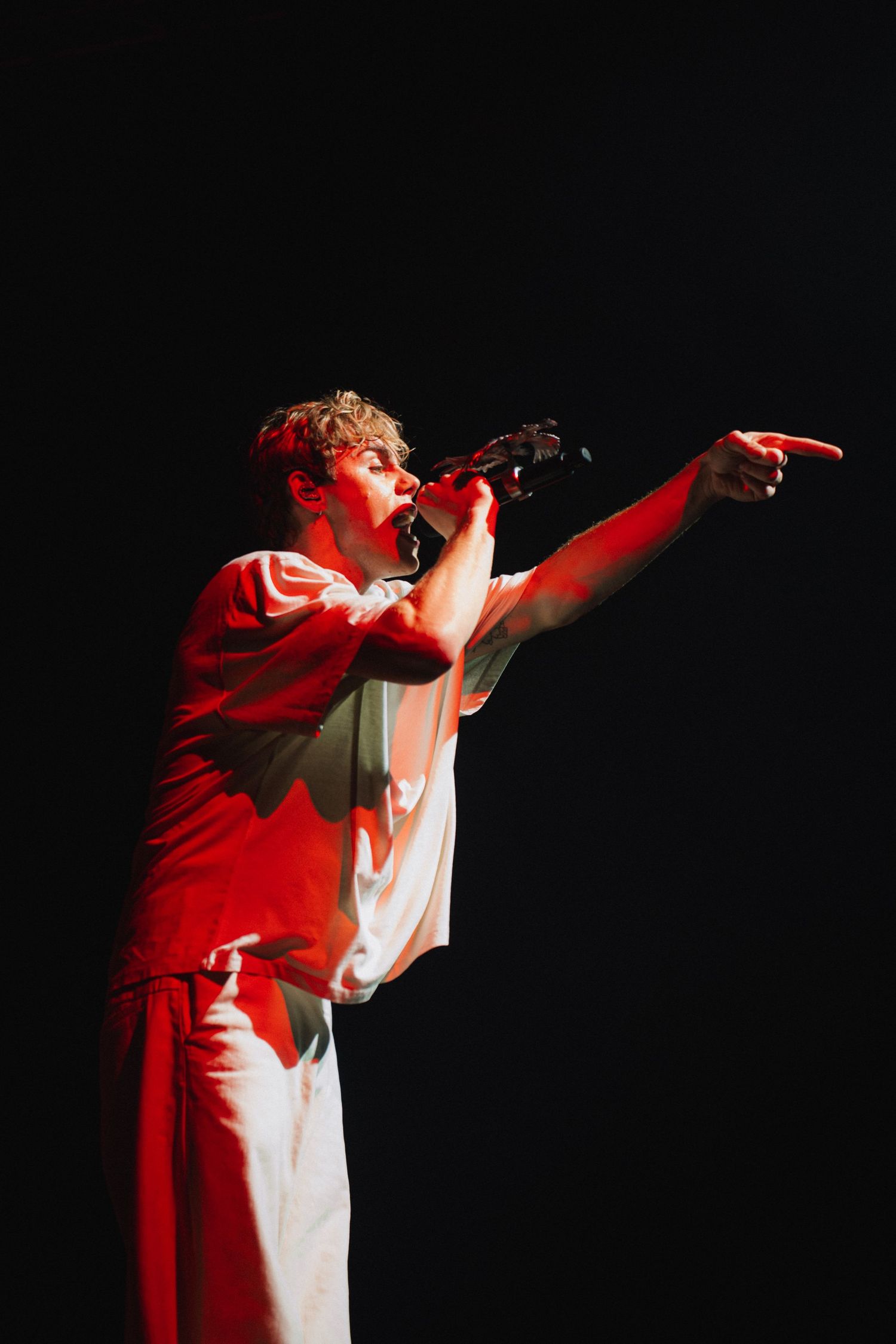 A performer in white clothing sings passionately on stage with dramatic red lighting and dynamic gestures.