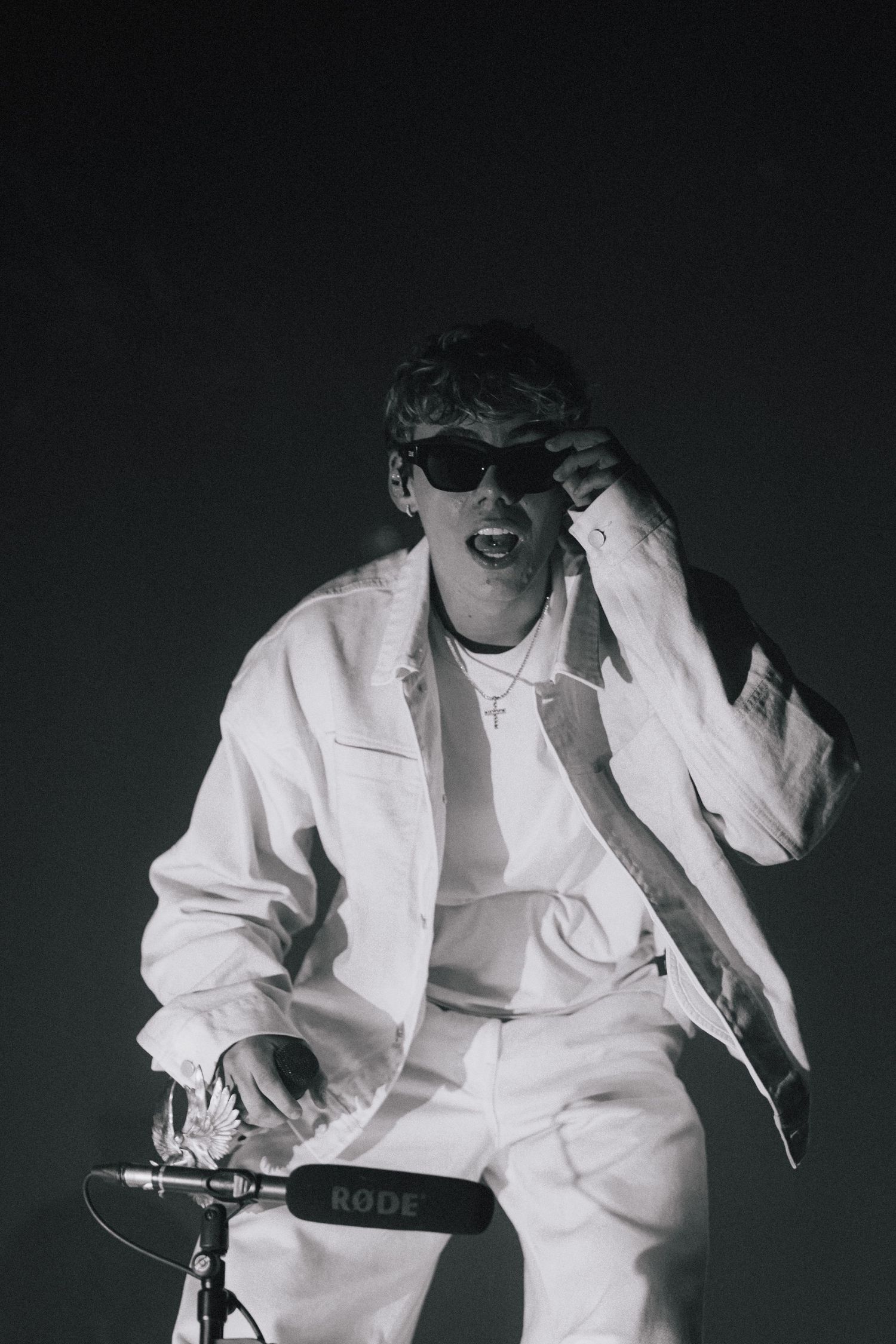 A person in all white clothing and sunglasses sits on a bike in a dramatic black and white photo against a dark background.
