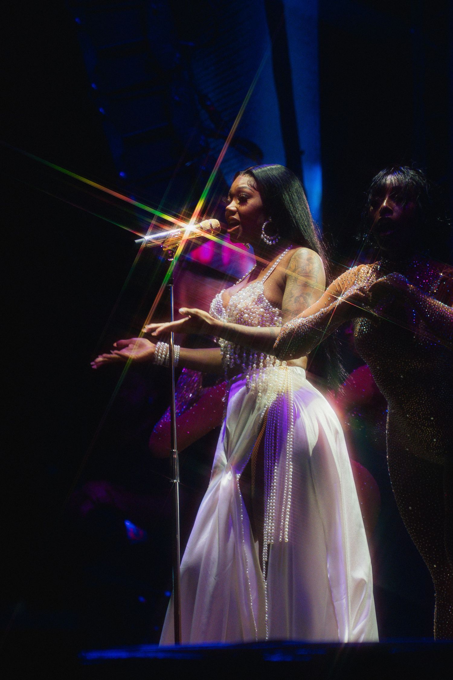 A performer in a flowing white dress sings on stage with dramatic lighting and sparkle effects in a dark theater.