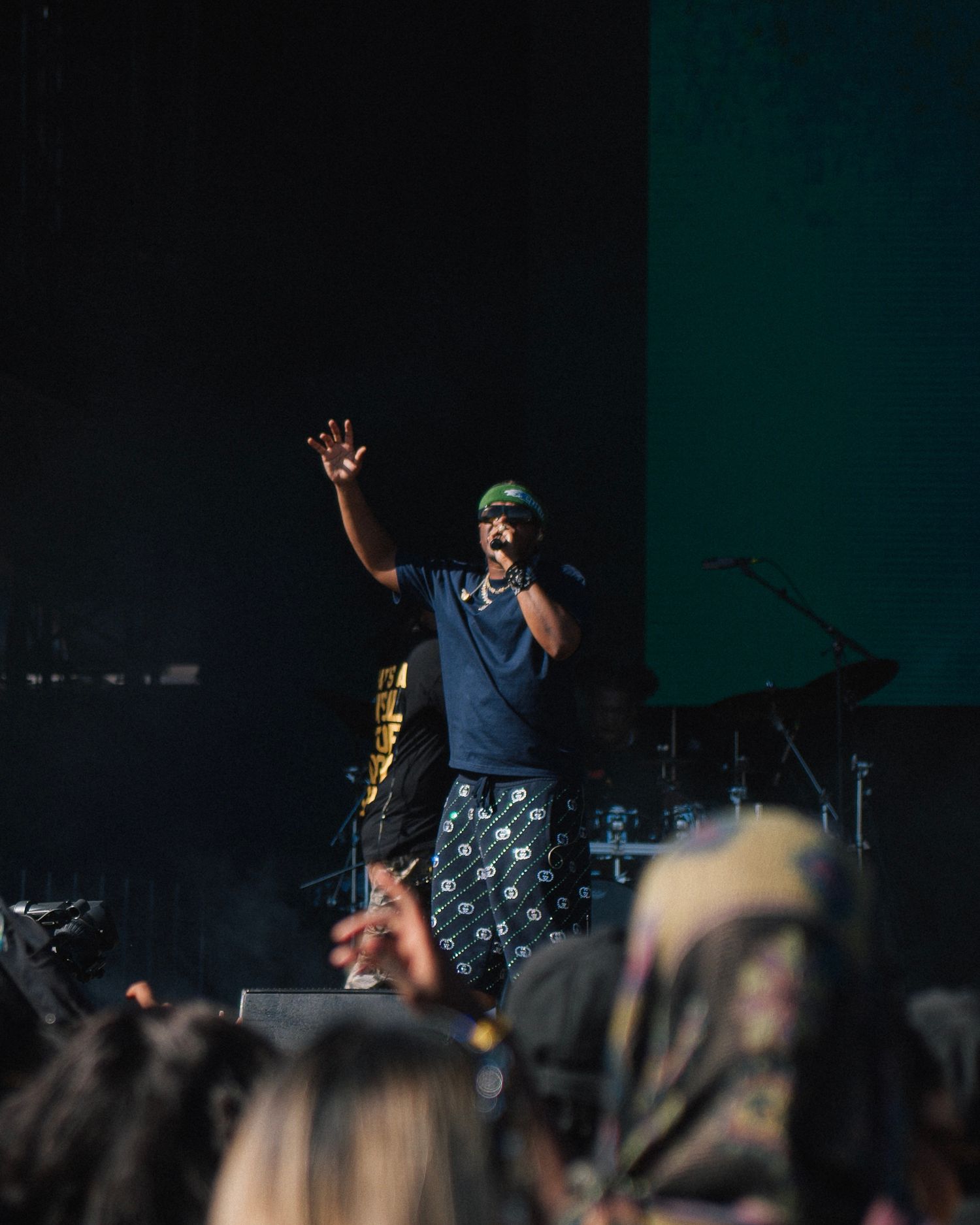 A performer in a cap and sleeveless shirt raises their arm on stage during a concert performance.