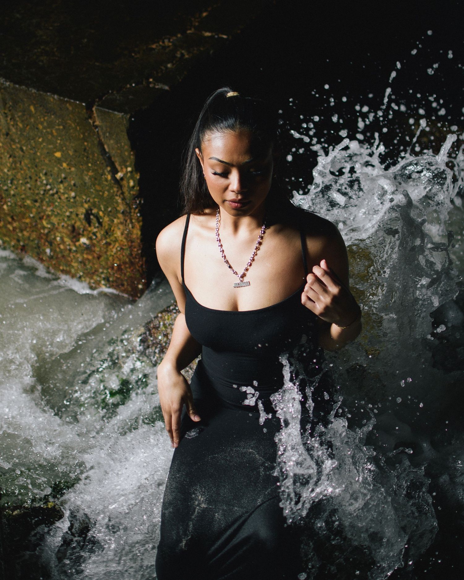 Figure in black dress poses against dramatic water splash on dark background with artistic lighting and beaded necklace.