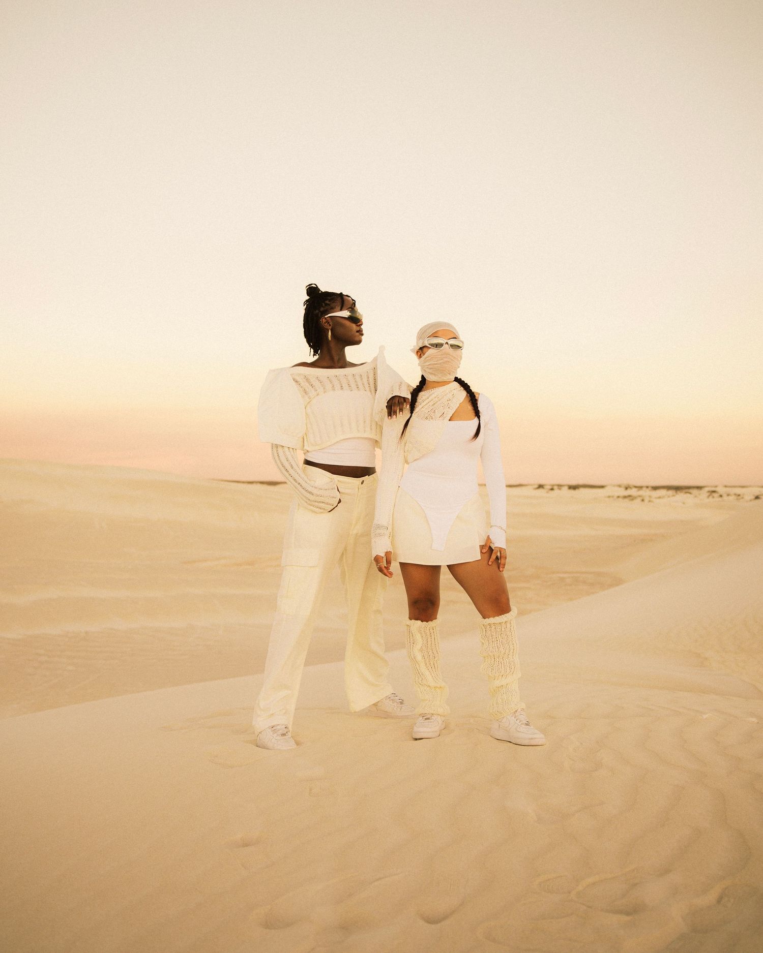 Two figures in white outfits posing against a desert sunset backdrop.