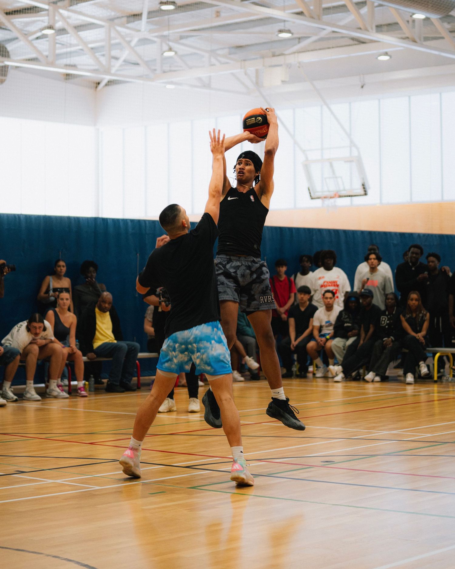 Indoor basketball game showing offensive player taking a jump shot over defender.