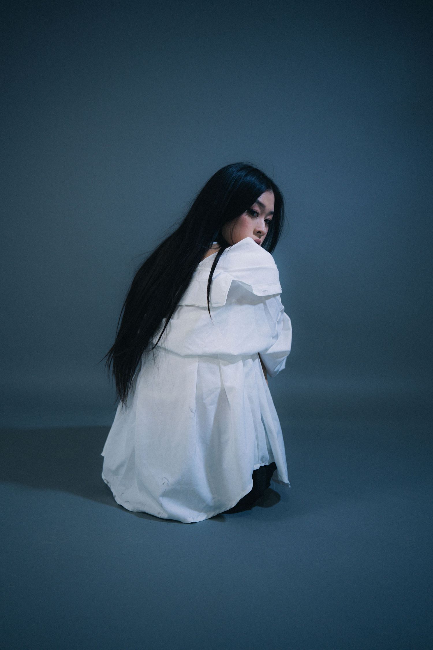 Figure in white dress crouched in contemplative pose against blue-gray backdrop.