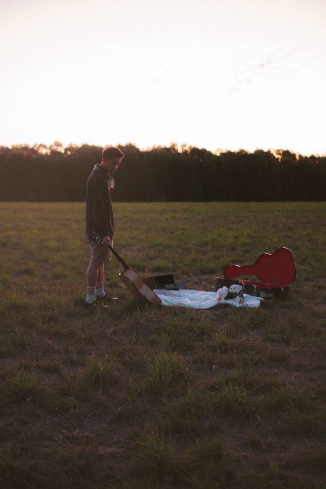 A silhouetted figure stands in a field at sunset with a picnic blanket and red guitar case on the ground.