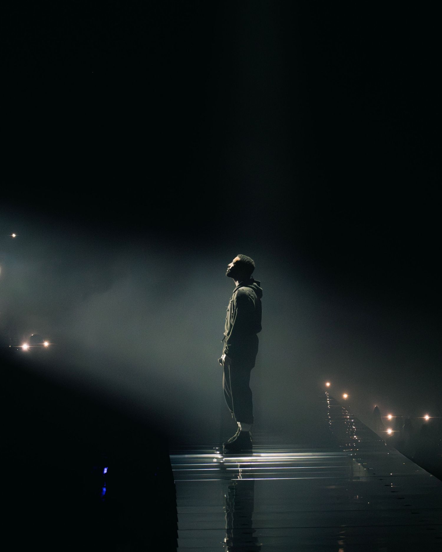 A dramatic silhouette stands on an elevated platform in fog with stage lights creating a moody atmosphere.