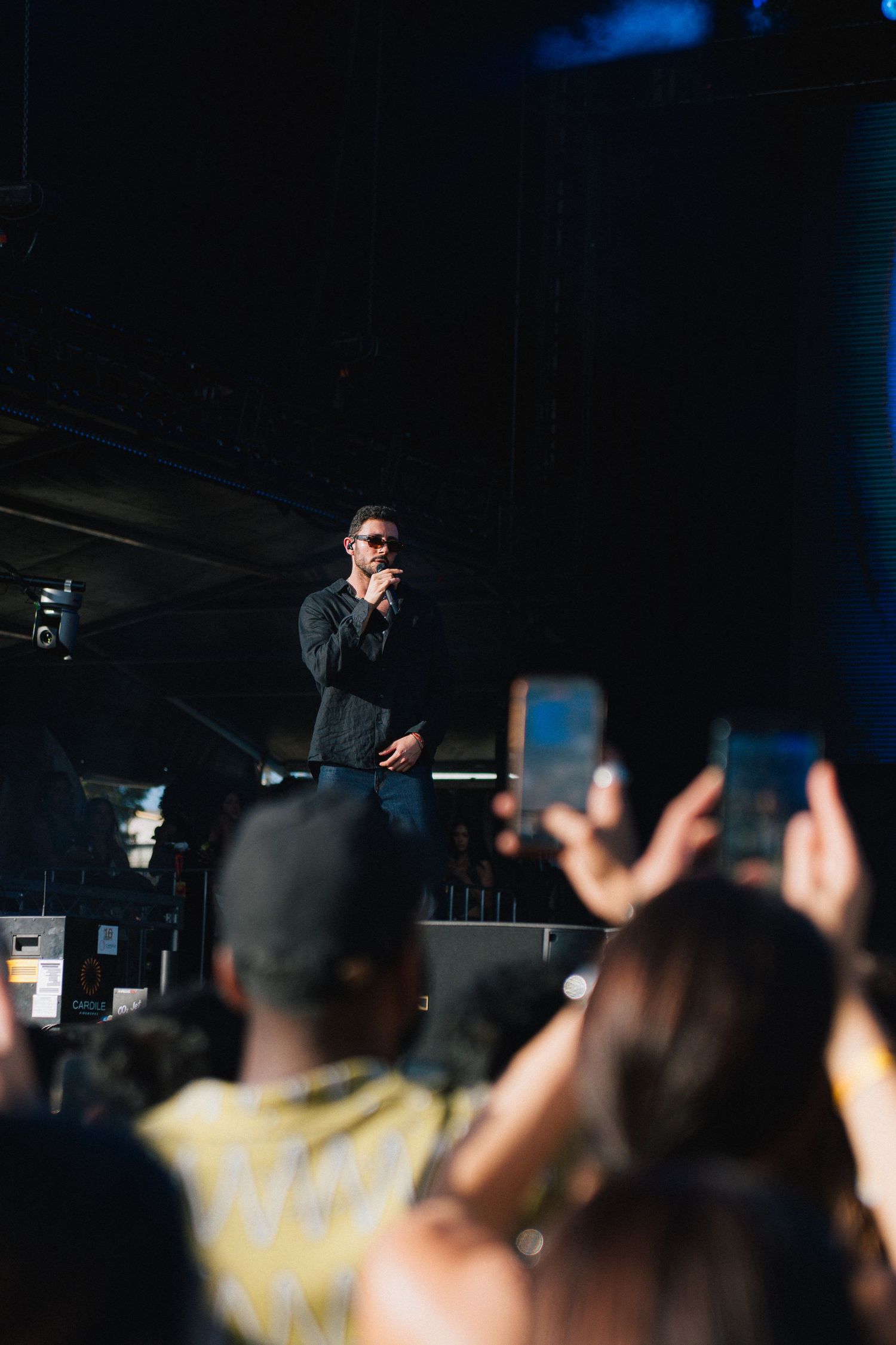 A performer on stage at a concert with audience members holding up phones to record in the dark venue.
