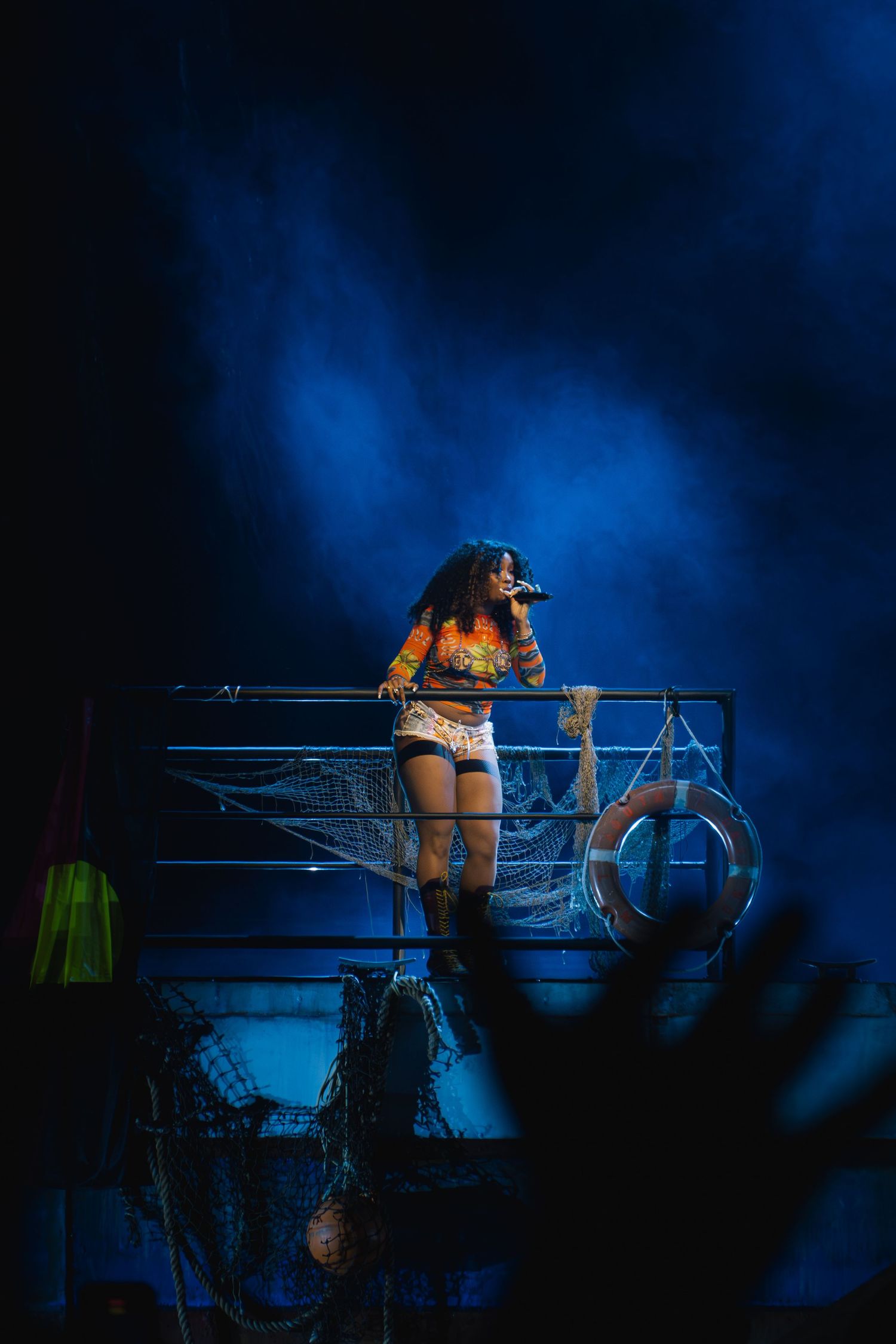 A performer in orange top and shorts stands on a stage platform with dramatic blue lighting and fog effects.