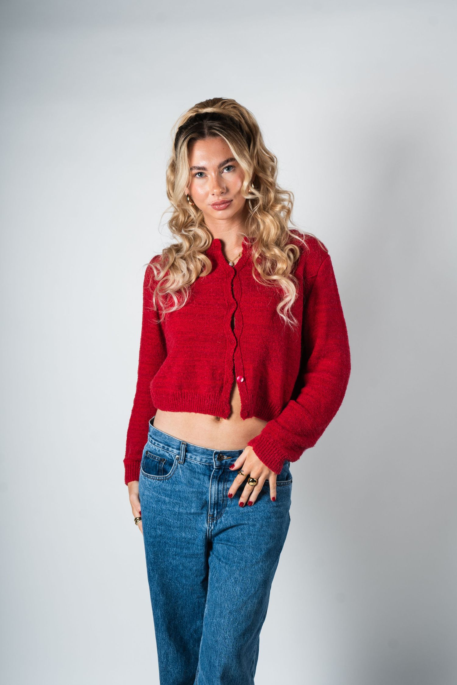 Person wearing a red cropped cardigan sweater and high-waisted blue jeans poses against white background.