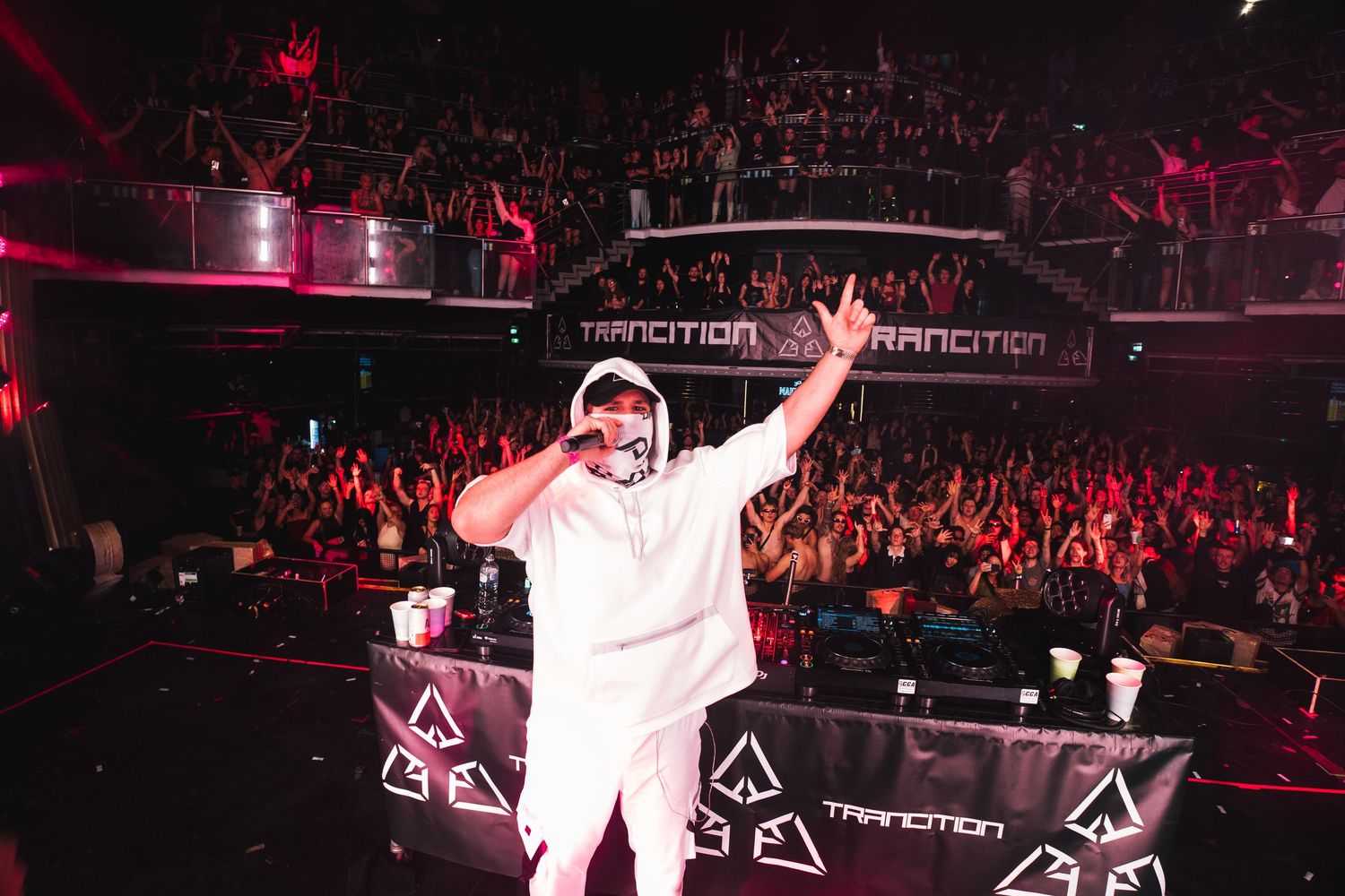 A performer in a white outfit silhouetted against a red-lit stadium crowd with multiple angled stage lights overhead.