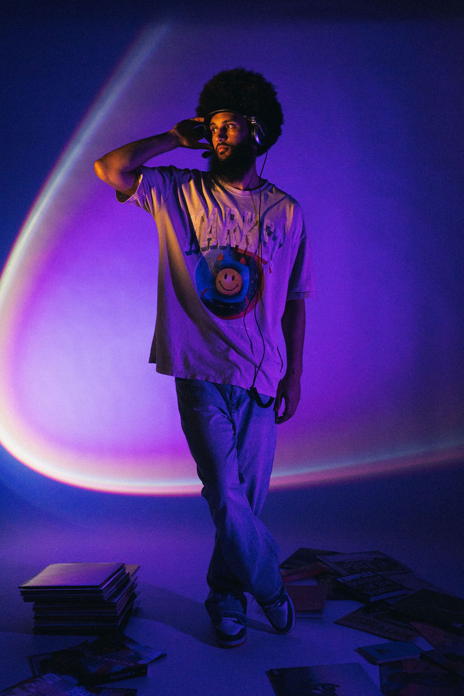 A person poses in casual wear with a smiley face graphic shirt against a purple and blue gradient background with light effects.