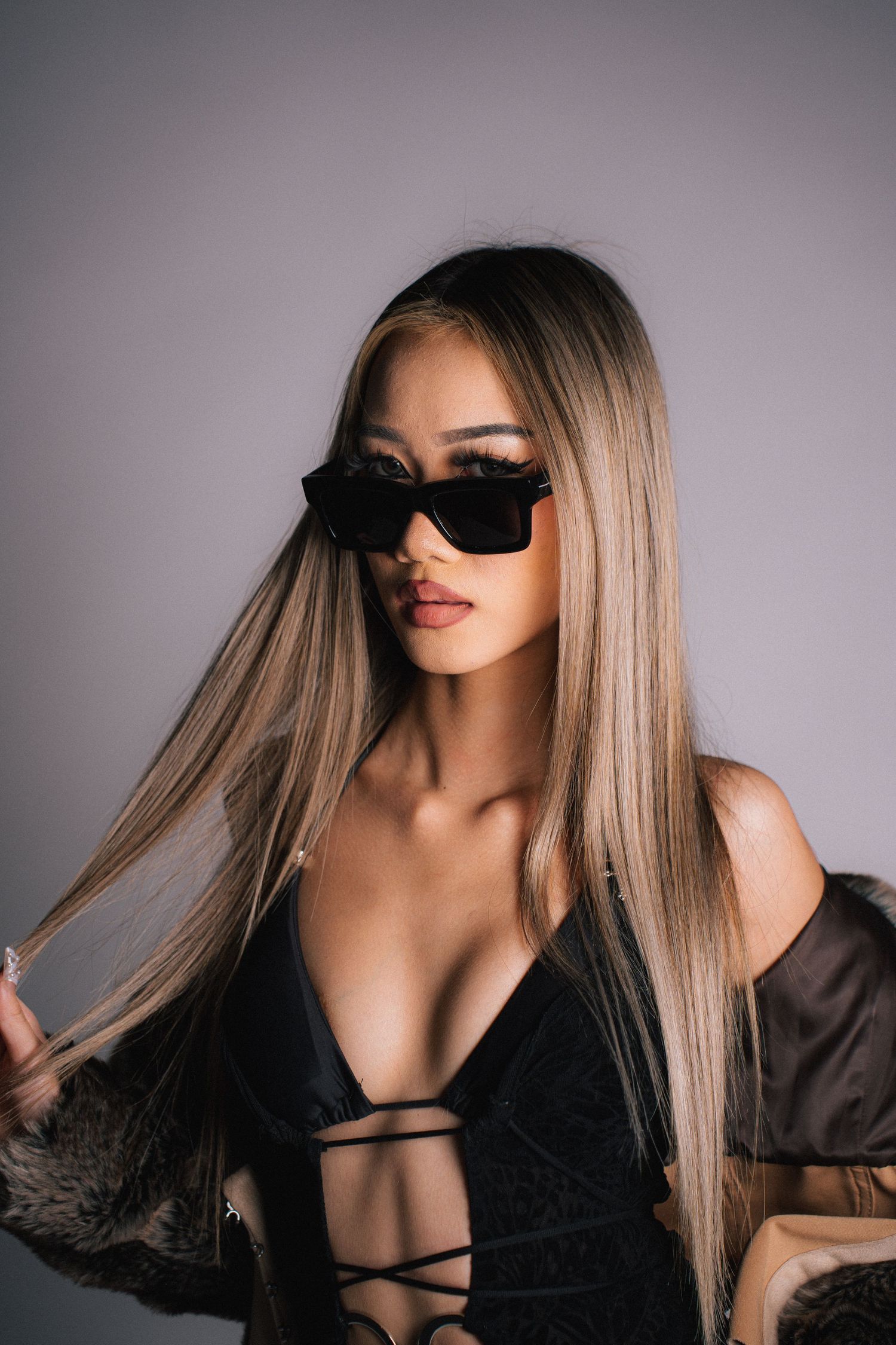 A figure with blonde hair poses against a gray backdrop wearing a black top and sunglasses in a moody fashion photoshoot.