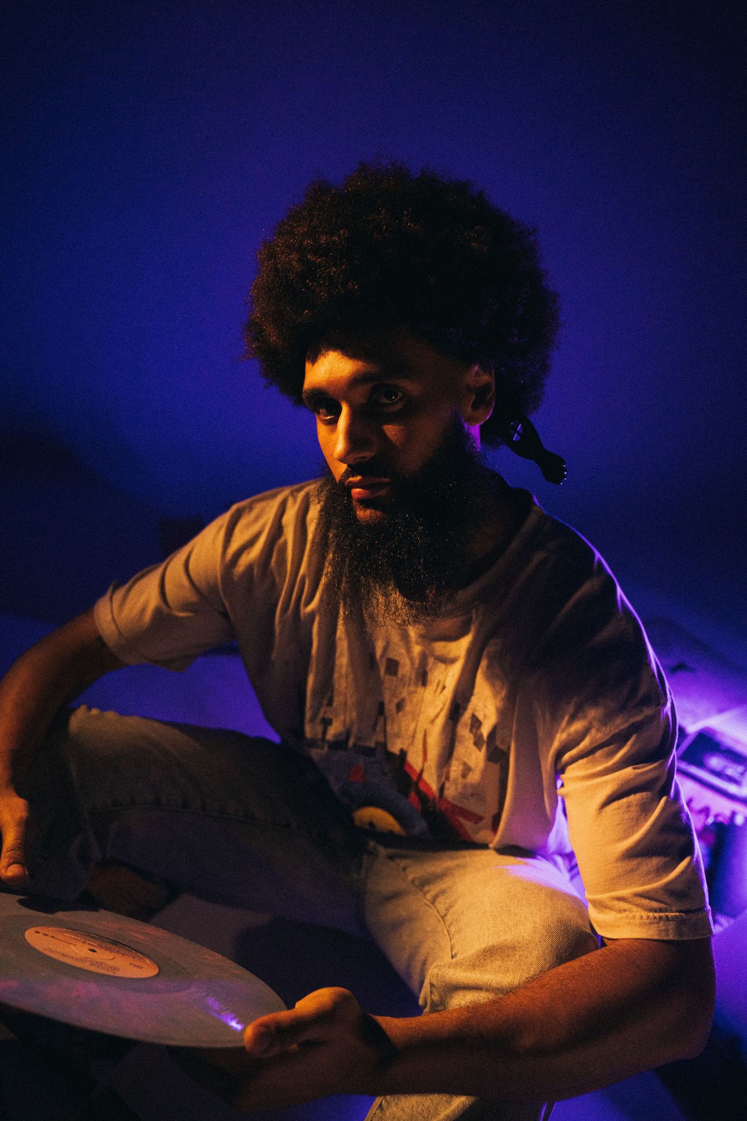 A DJ in a white shirt performs at a club with purple lighting illuminating the background, focused on mixing tracks.