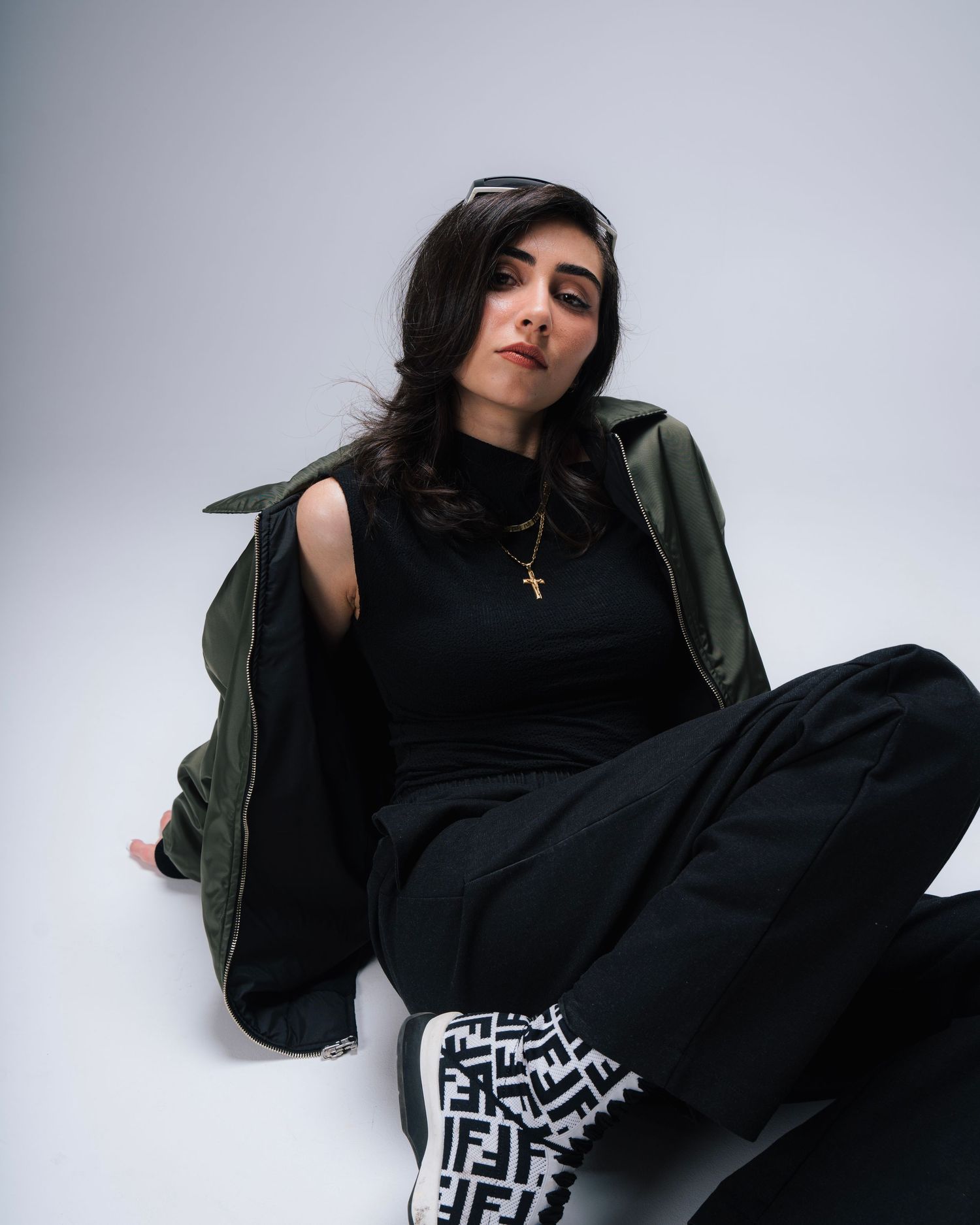 Stylish portrait against white backdrop showing figure in black outfit with olive jacket and patterned pants.