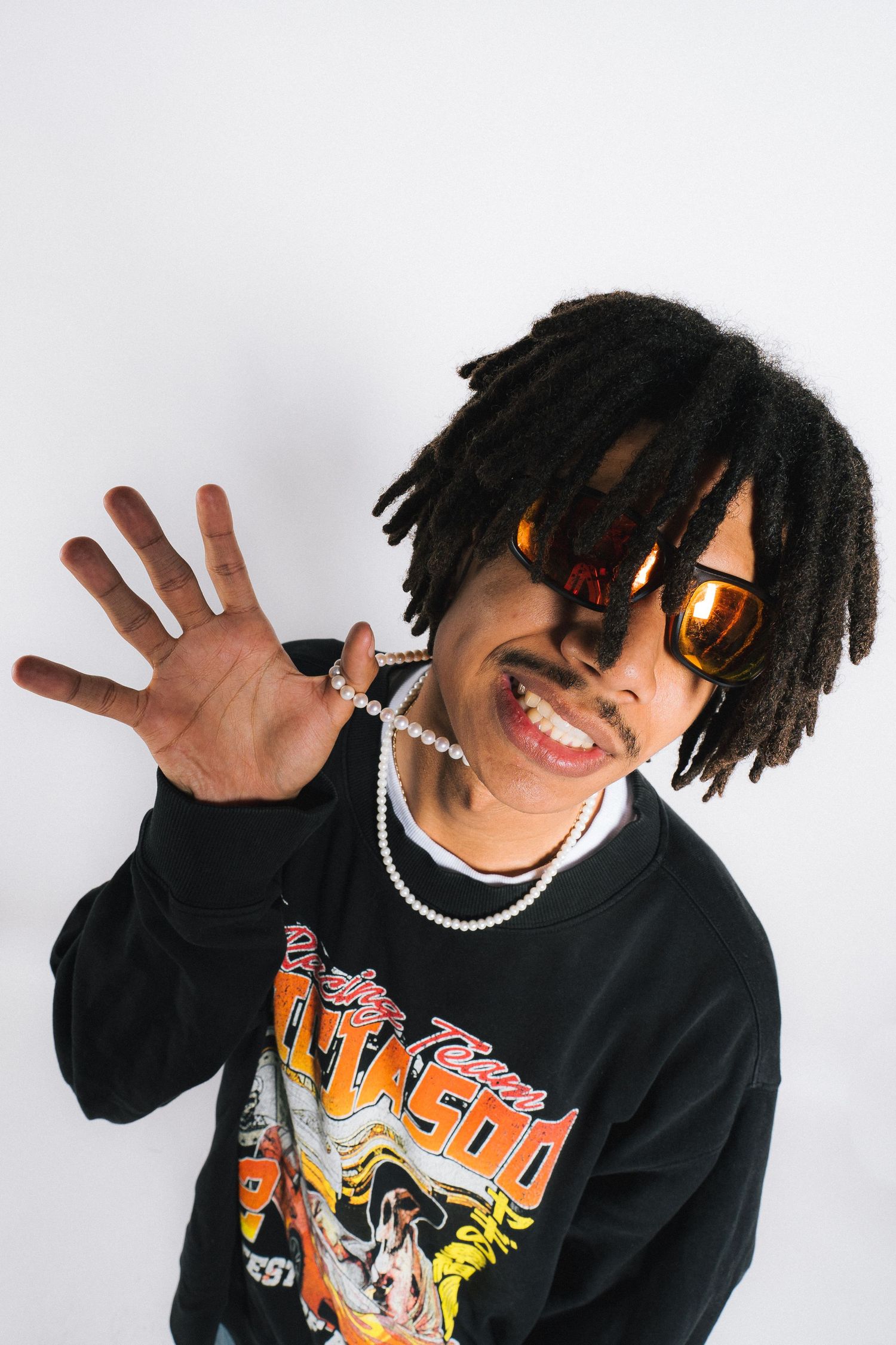 Expressive person wearing sunglasses and black anime graphic sweatshirt poses with raised hand against white background.
