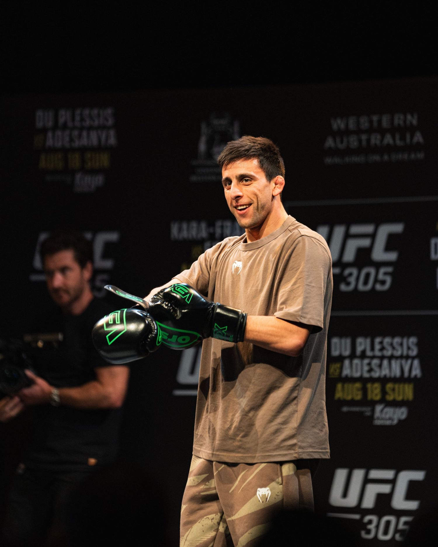 Fighter smiling while holding green UFC gloves during promotional event.