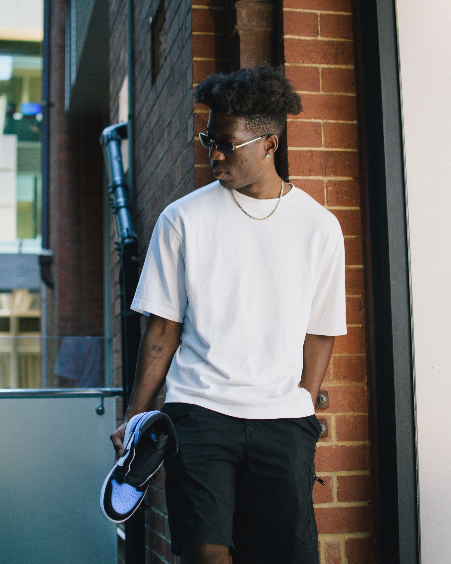 Stylish person in white t-shirt and shorts stands against brick wall wearing sunglasses.