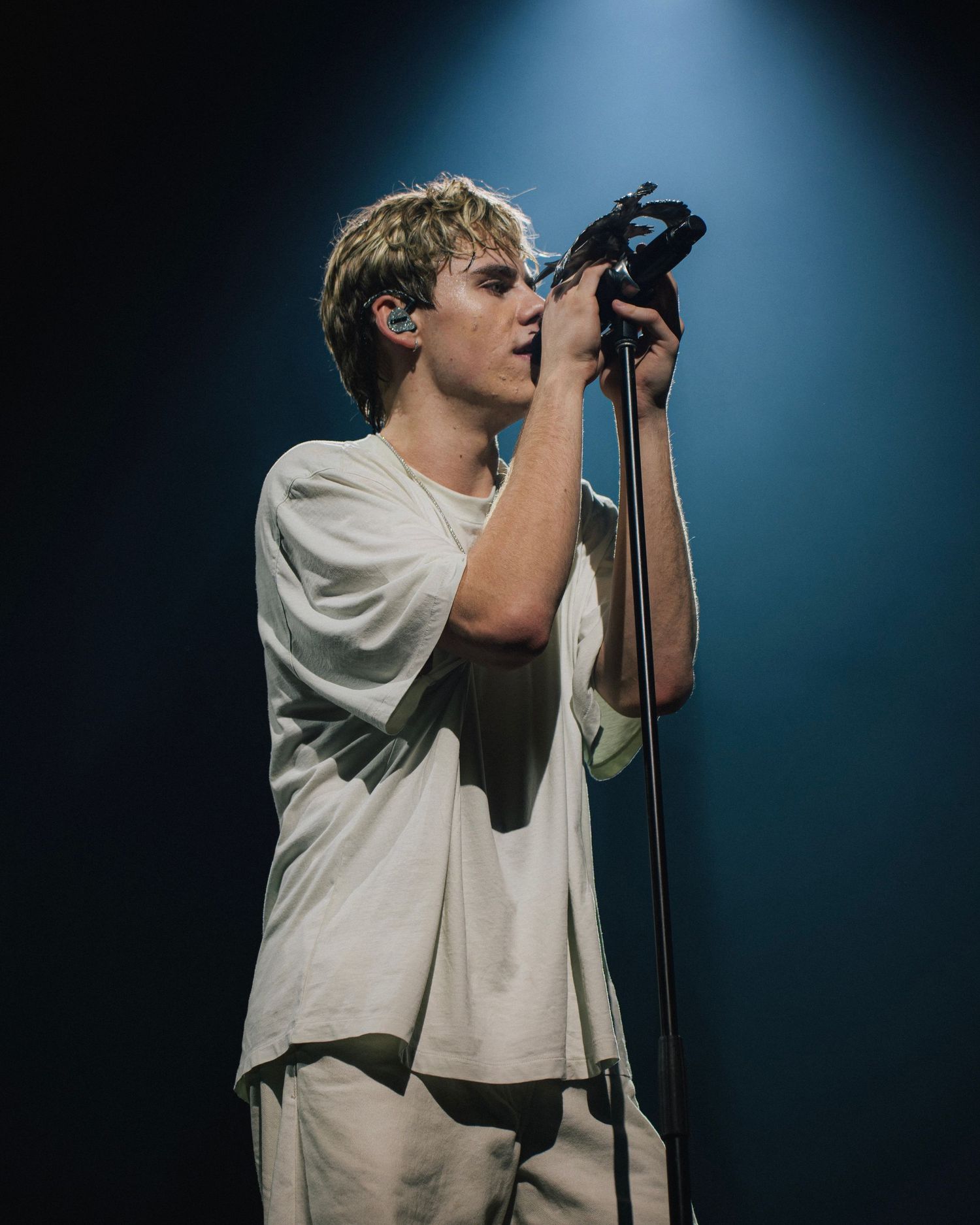 A performer wearing a white shirt sings into a microphone on stage with dramatic blue lighting in the background.