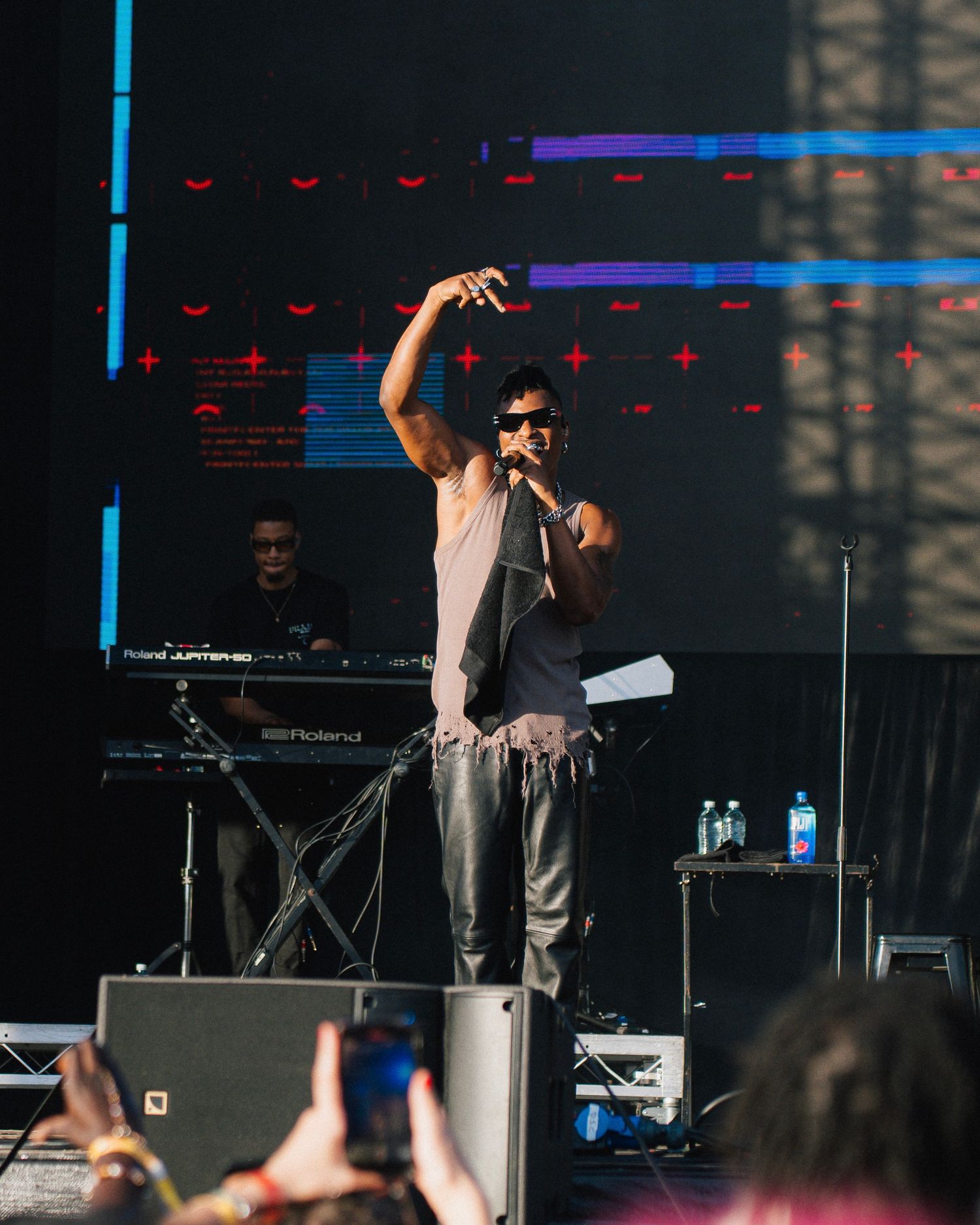 Performer in sunglasses and casual attire raises arm while singing on stage with electronic equipment and lights in background.