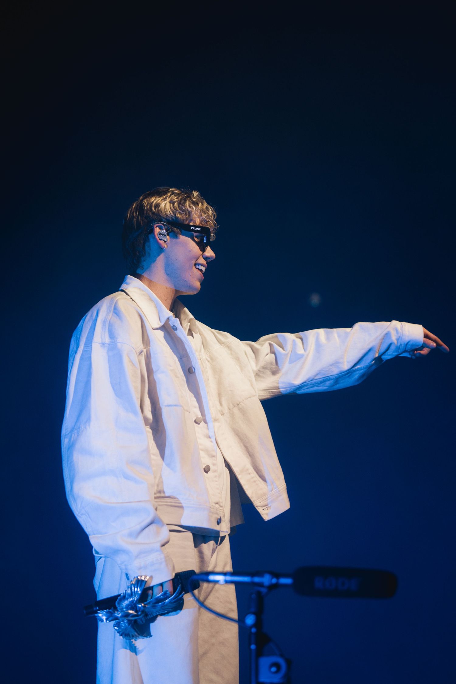 Performer in white outfit and glasses gestures dramatically on stage against dark blue background during concert performance.