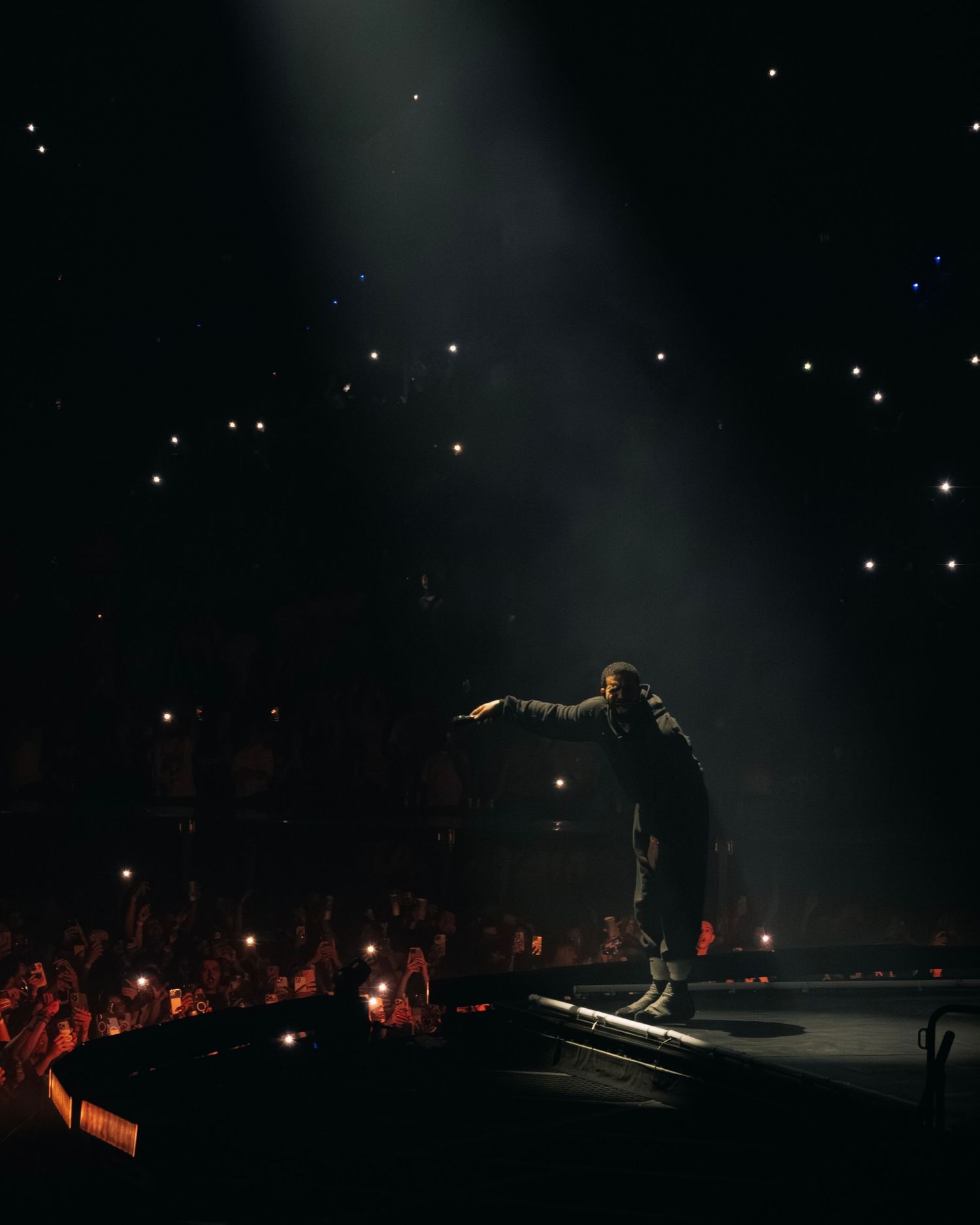 Silhouetted performer stands on stage under dramatic spotlight beam with crowd visible below at nighttime concert.