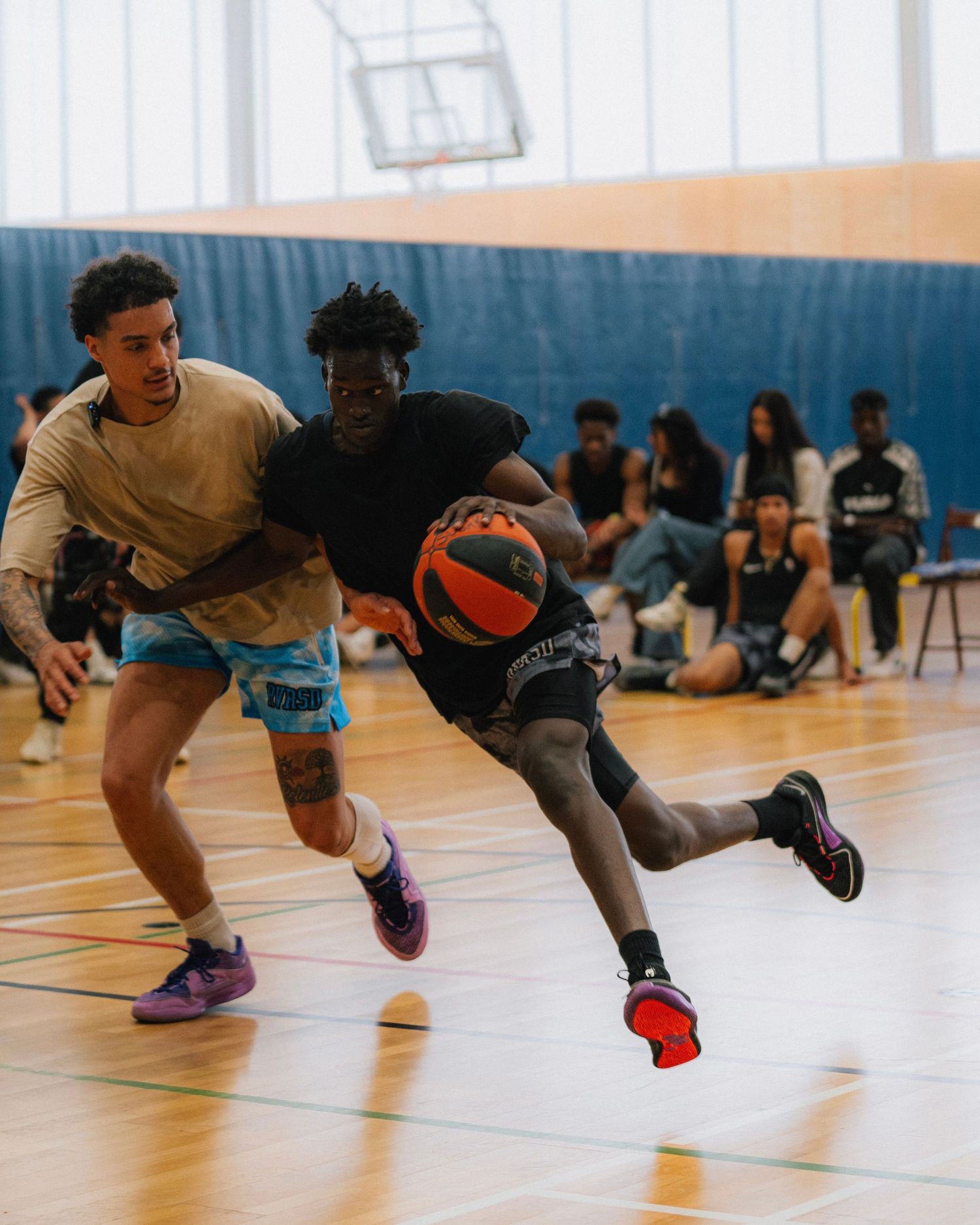 Two players engage in an intense basketball dribble drive on a gymnasium court.