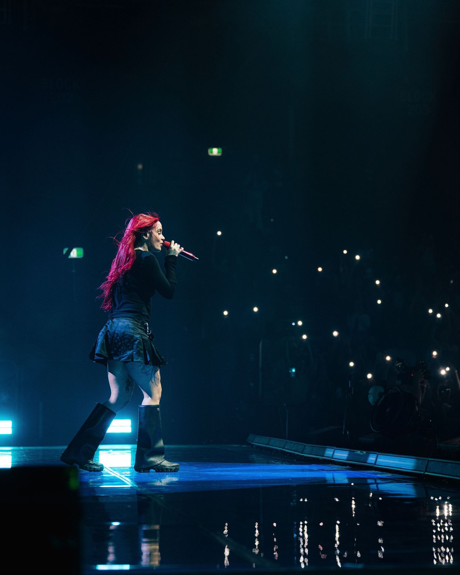 Performer with bright pink hair singing on stage under blue lighting with glowing phone lights in the dark audience background.