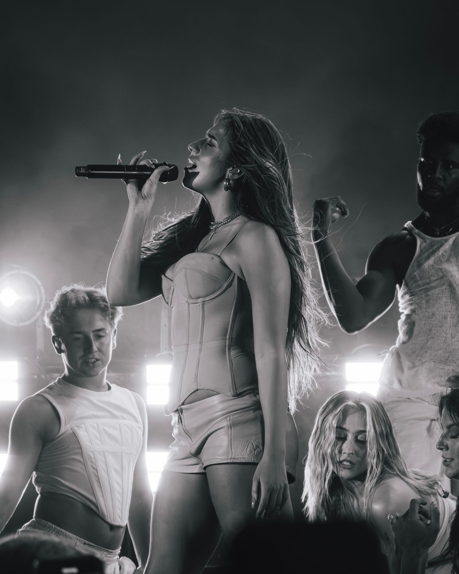 A concert performance in black and white with performers on stage in white outfits under dramatic spotlights.