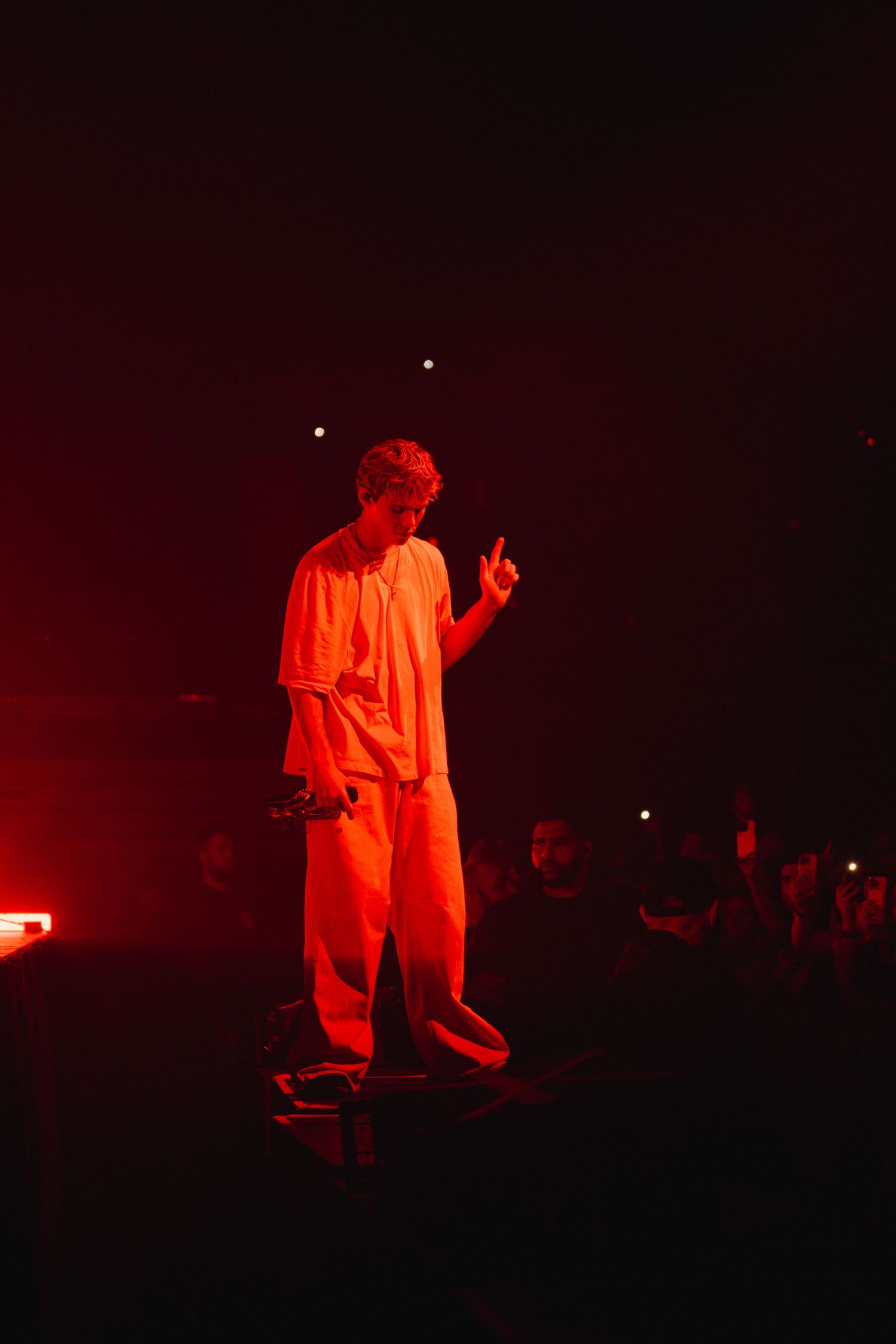 A performer in red lighting stands on stage with one finger raised during a dramatic concert performance.