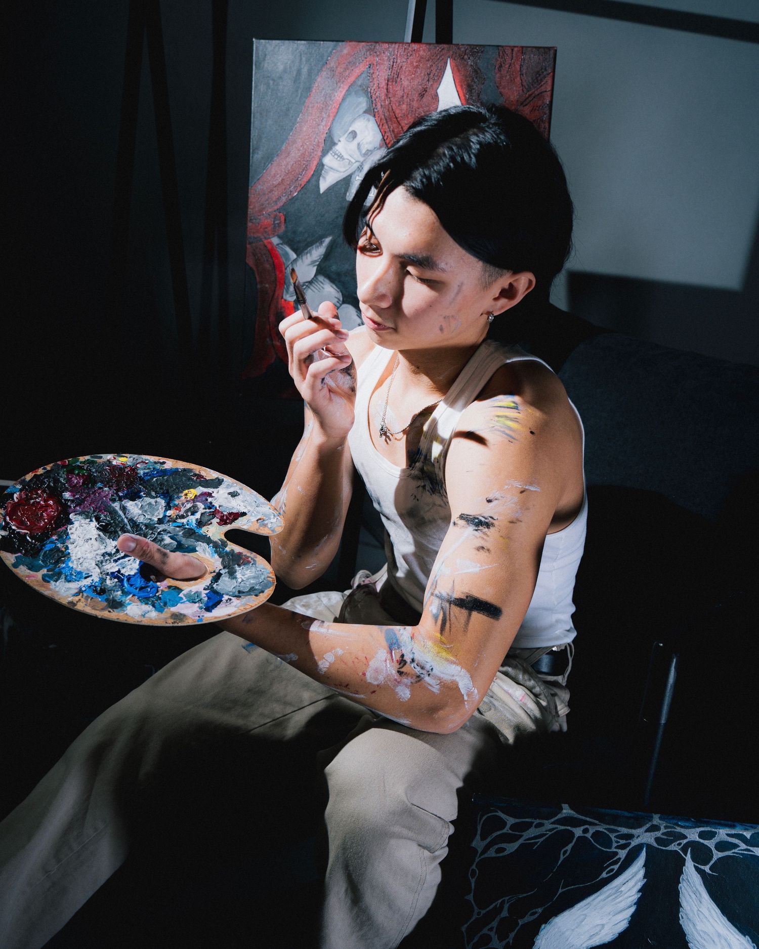 Artist holds paint palette while working on colorful paintings in dimly lit studio.