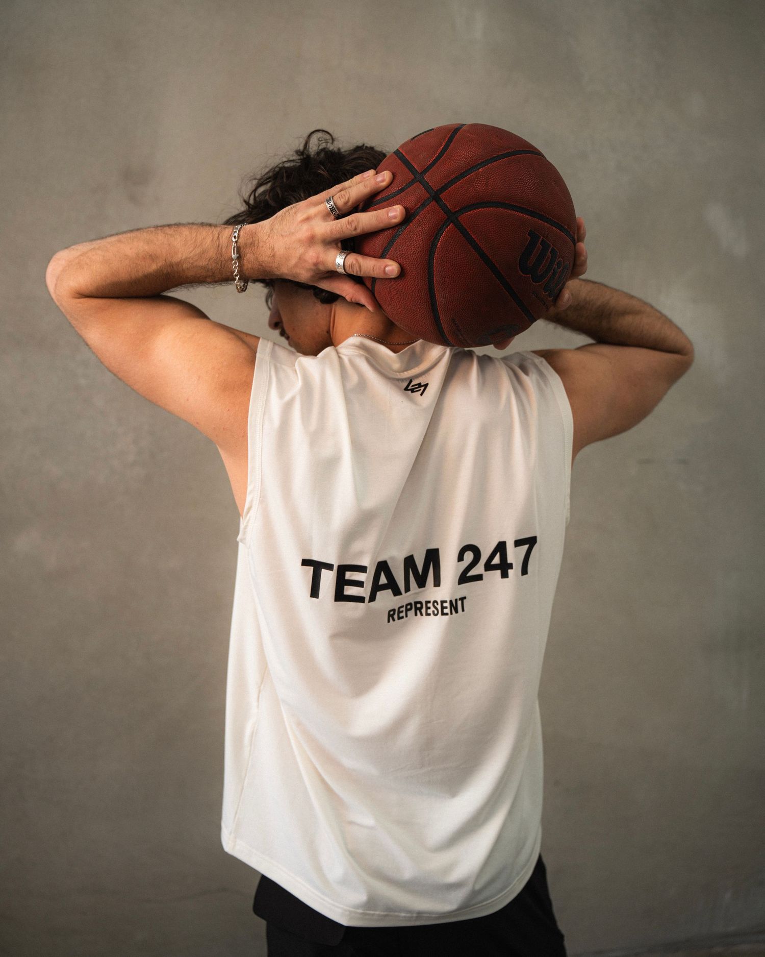 Person wearing Team 247 white t-shirt holds basketball behind their head against gray wall.
