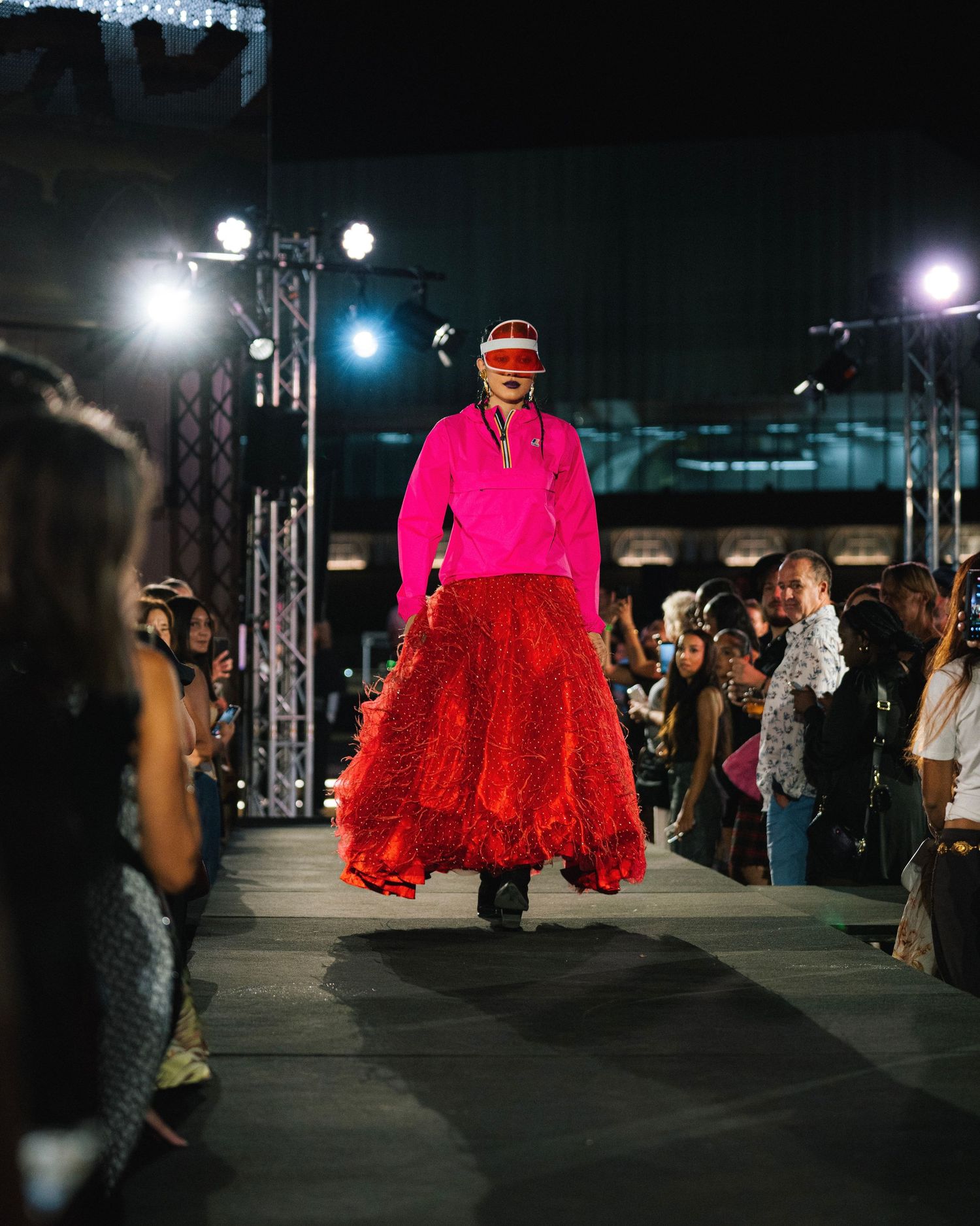 Model walks runway in striking hot pink top and flowing red skirt during fashion show.