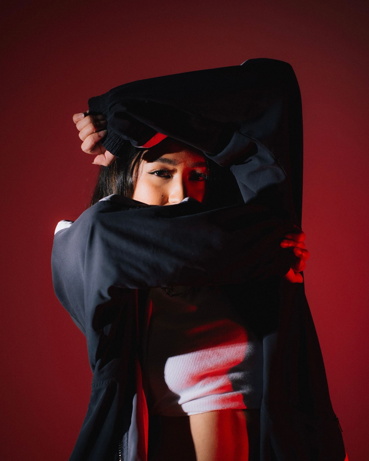 Figure in dark clothing partially covering face against red background with dramatic lighting.