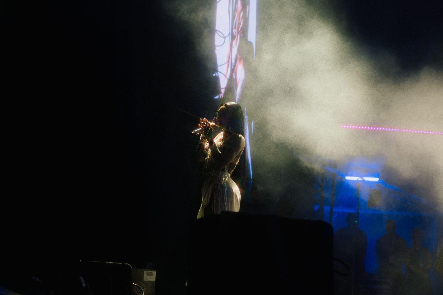 A silhouetted performer stands on a dark stage with dramatic beams of light and fog effects in the background.