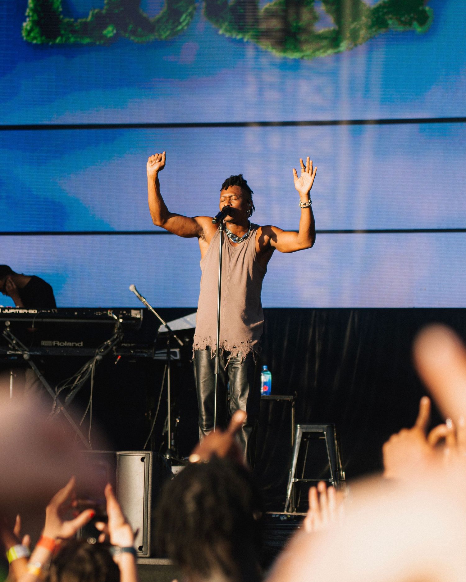 Performance artist raises arms on stage with keyboard instruments and audience silhouettes in foreground against blue backdrop.