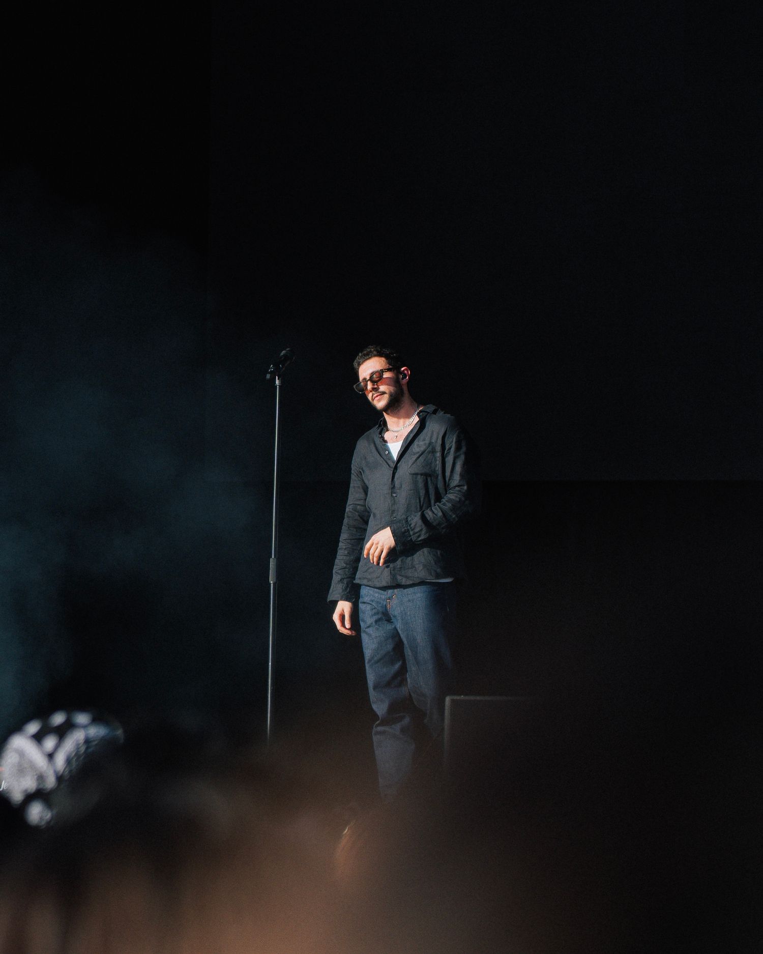 A performer stands on a dimly lit stage with atmospheric blue lighting and a microphone stand in the foreground.