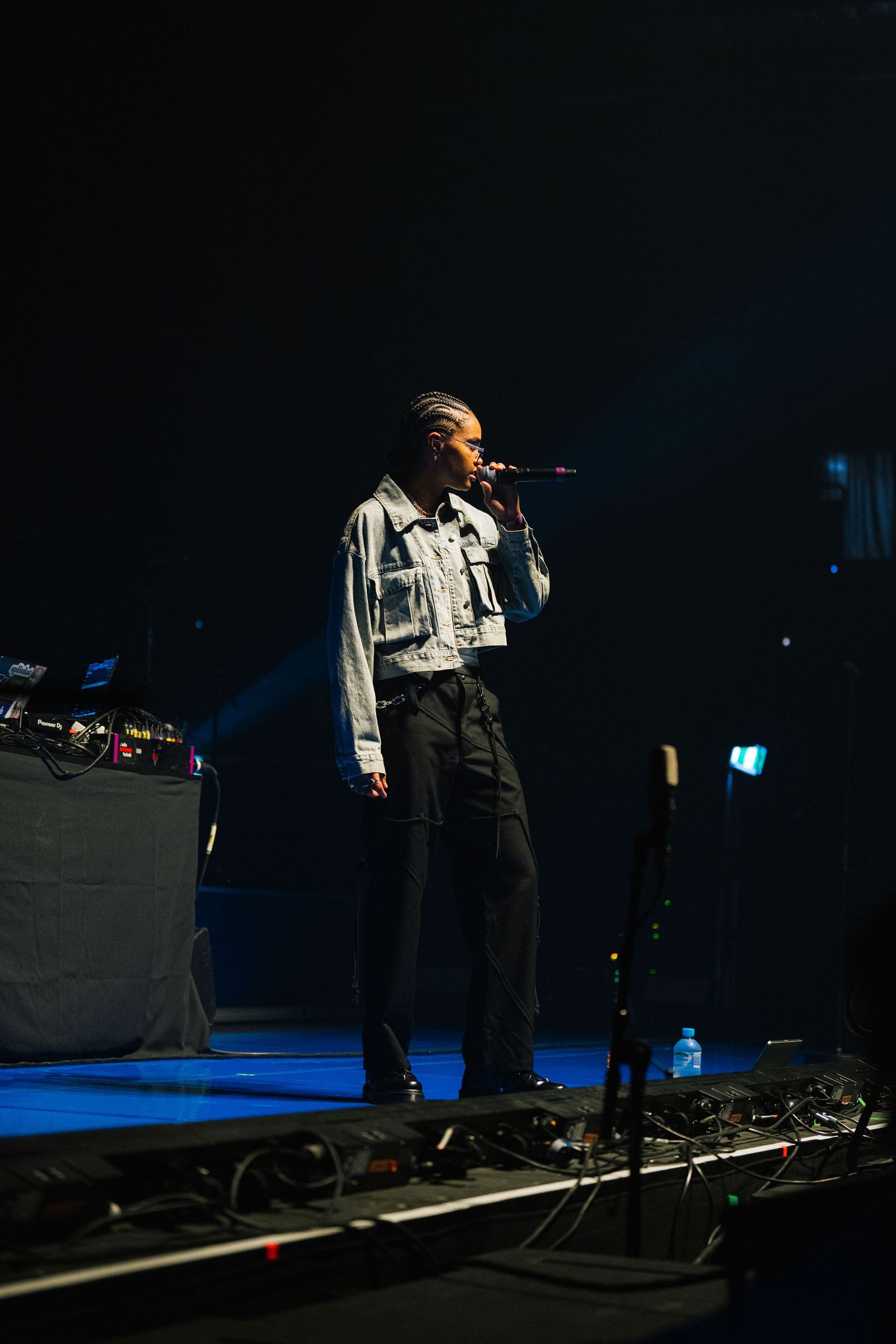 A performer in a light denim jacket and dark pants sings on stage with blue lighting and audio equipment visible.