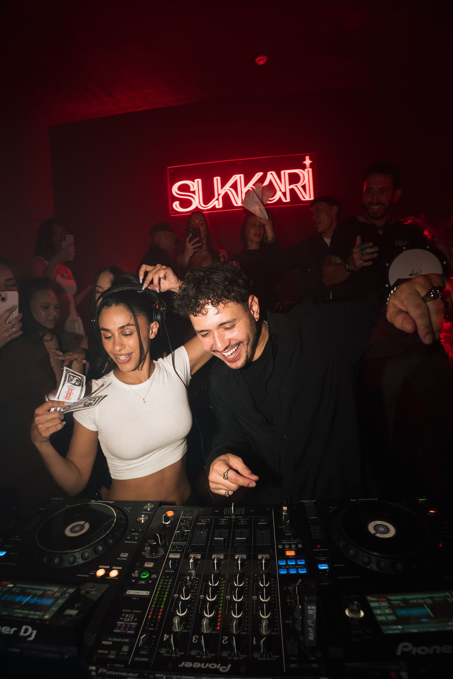 DJs perform at a nightclub with red neon 'SUKARI' sign glowing in the background while partygoers dance and celebrate.