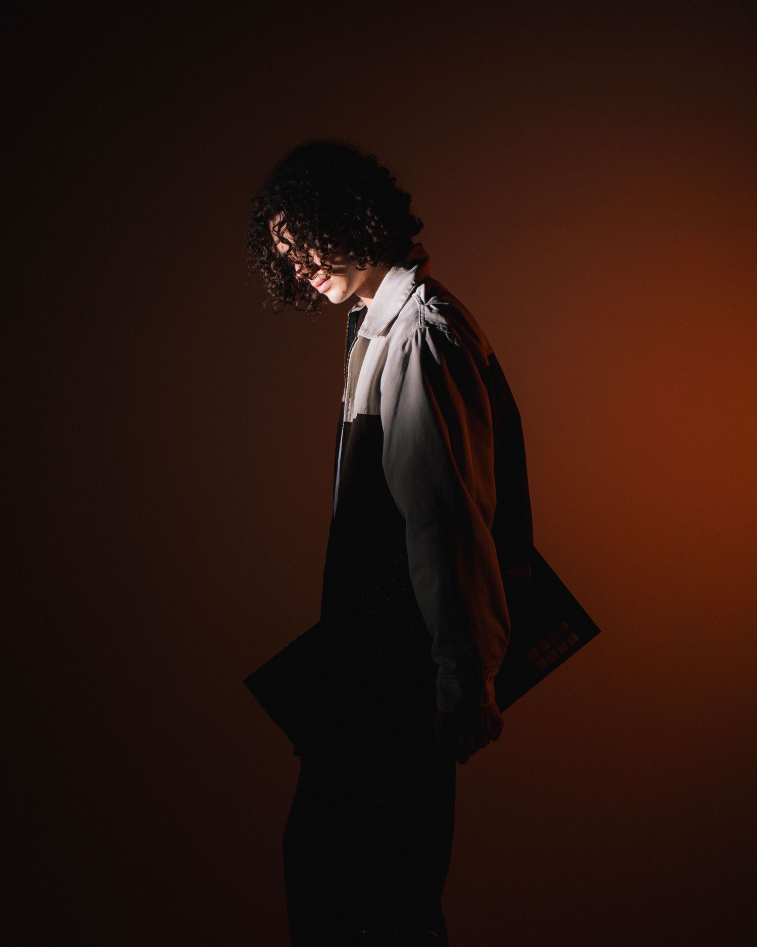 Silhouette figure with curly hair in formal attire poses dramatically against dark reddish-brown backdrop.